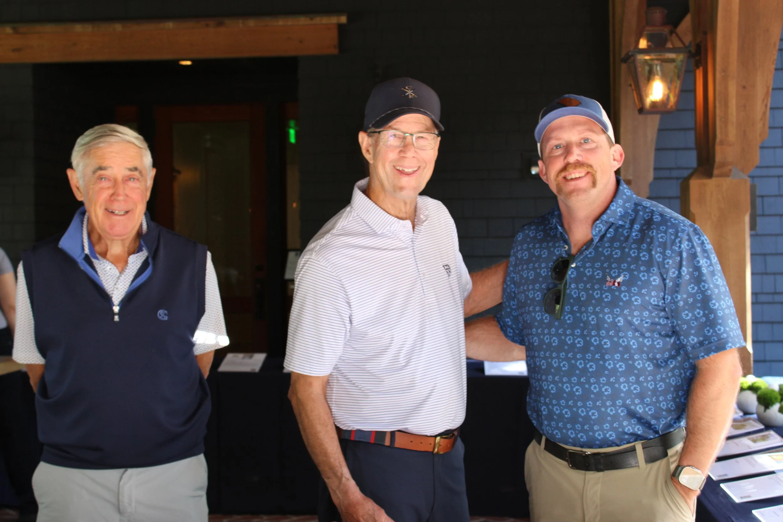 Three middle-aged men smiling at a social event indoors, standing side by side in casual and semi-formal attire, with wooden decor and a lantern-style light fixture in the background.