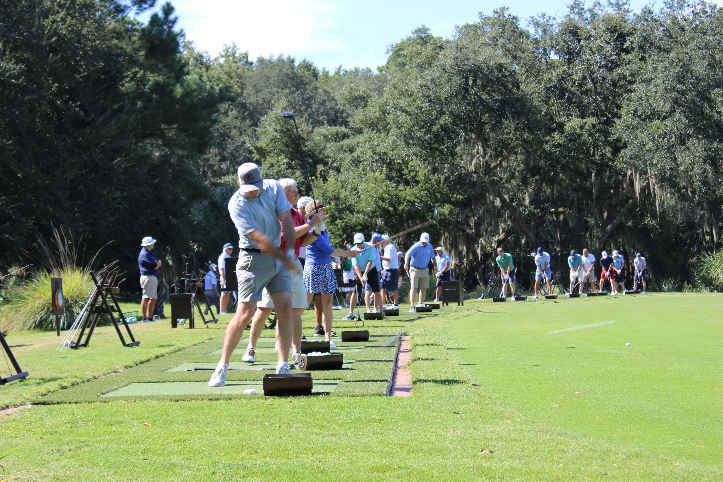 People practicing golf at an outdoor driving range surrounded by trees.