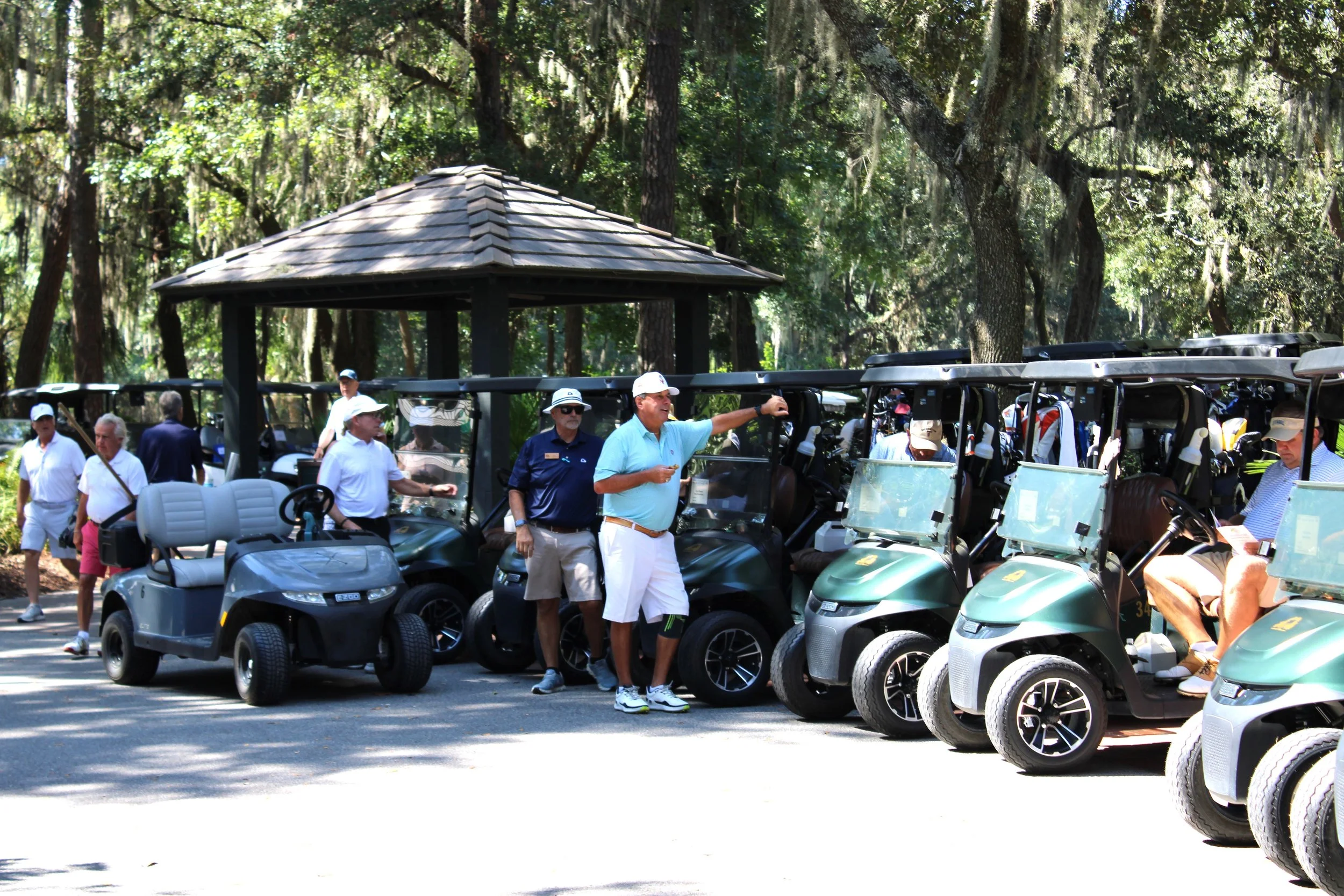 A group of people standing next to golf carts in a wooded area, with tall trees and sunlight filtering through the leaves.