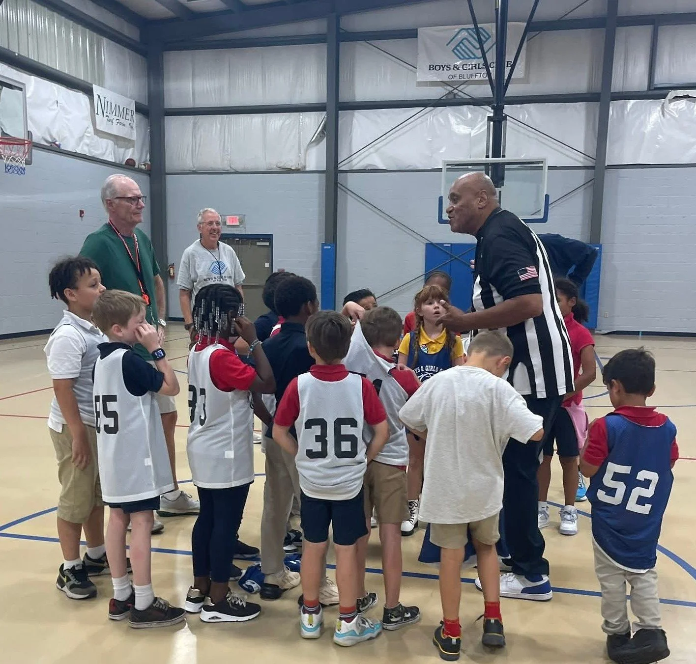 A group of young children wearing basketball jerseys listening to a referee speaking to them inside a gymnasium. Three adults are standing nearby, with one smiling, as the children gather around the referee.
