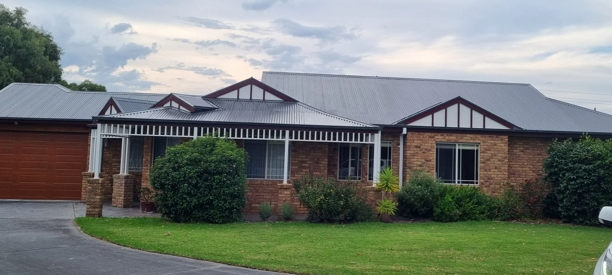 A brick house with a metal roof, a covered front porch with white posts, and a front yard with green grass, bushes, and trees under a partly cloudy sky.
