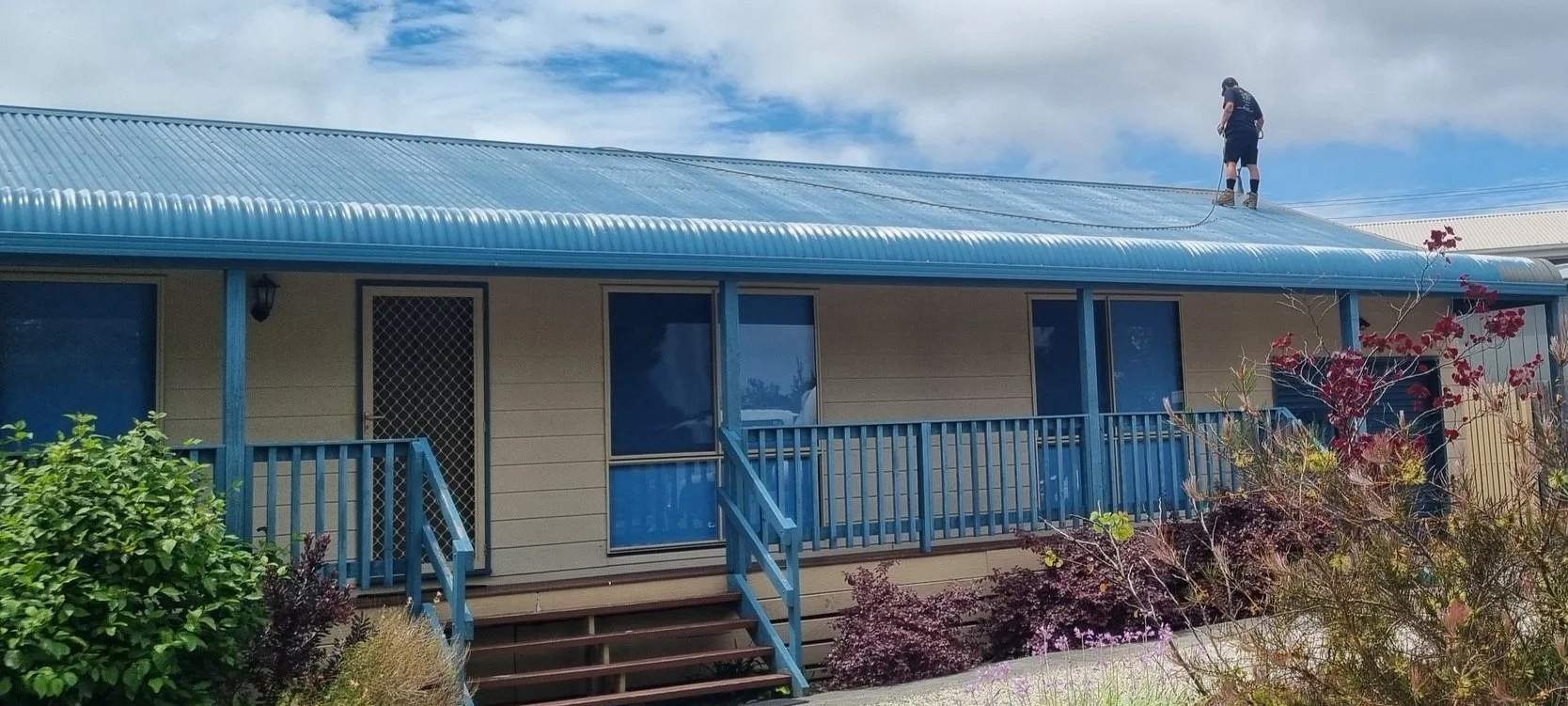 A person standing on a blue metal roof with safety harnesses, working on cleaning or maintenance during daytime. The house has beige siding, blue trim, and a small front porch with stairs surrounded by shrubs and plants. The sky is partly cloudy.