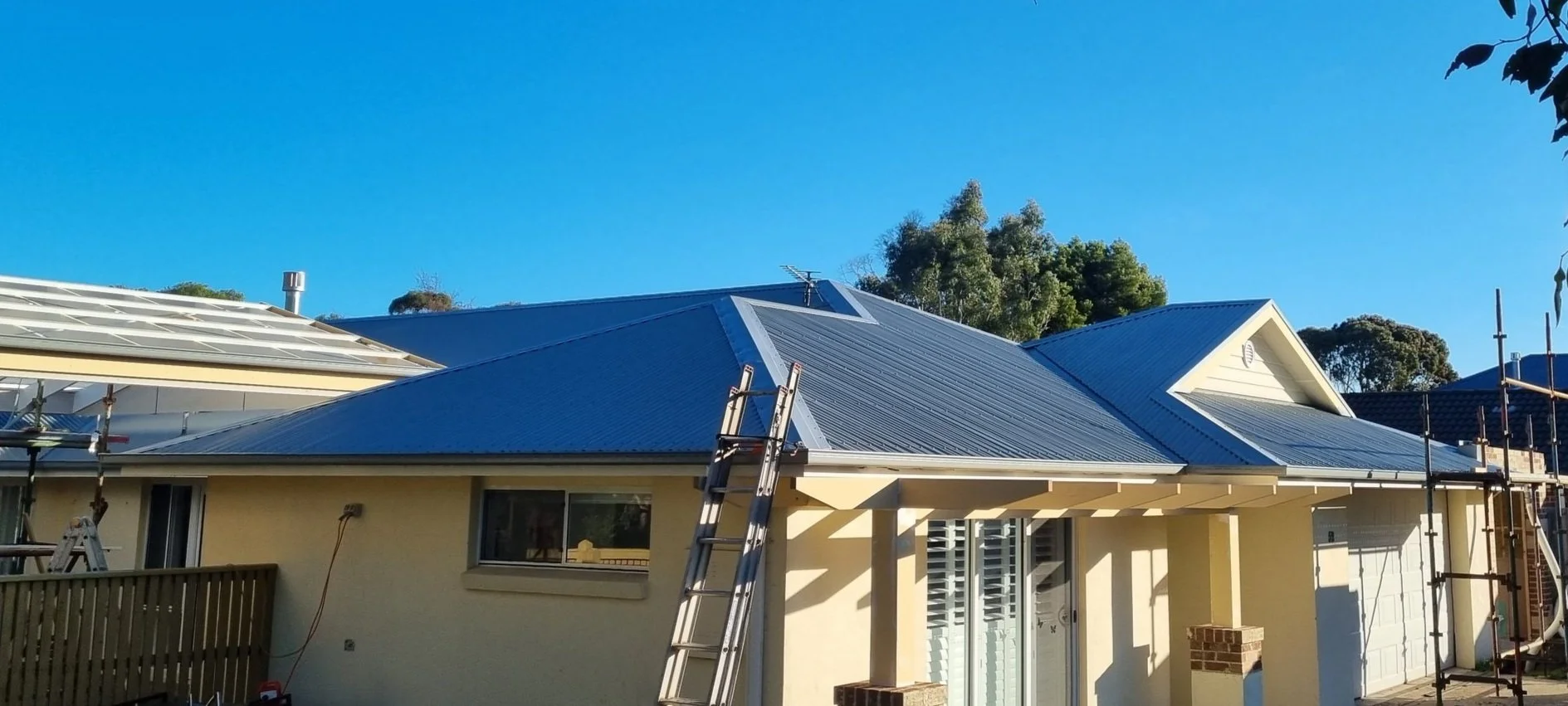 Residential house with a metal roof under construction, ladder leaning against the roof, scaffolding on one side, clear blue sky, and trees in the background.