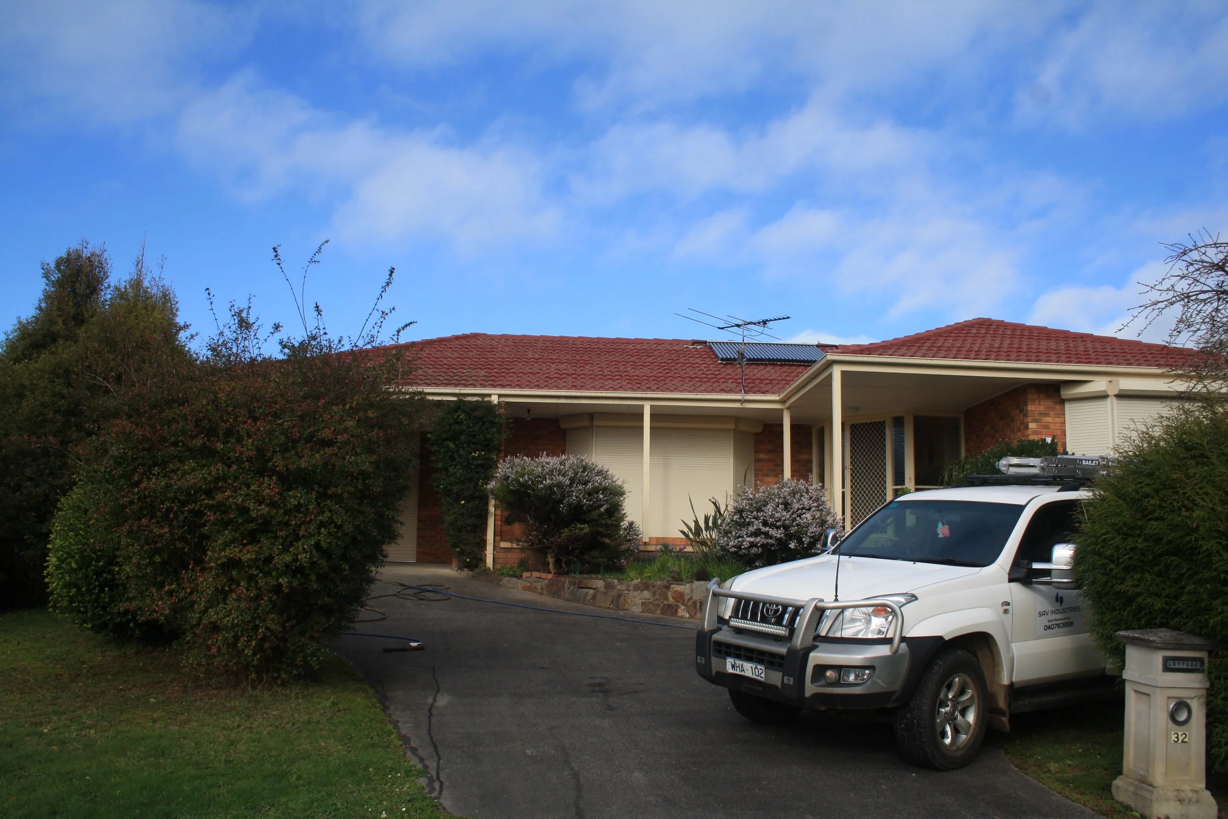 Single-story brick house with a red tiled roof, surrounded by bushes and trees, and a white utility vehicle parked on the driveway.