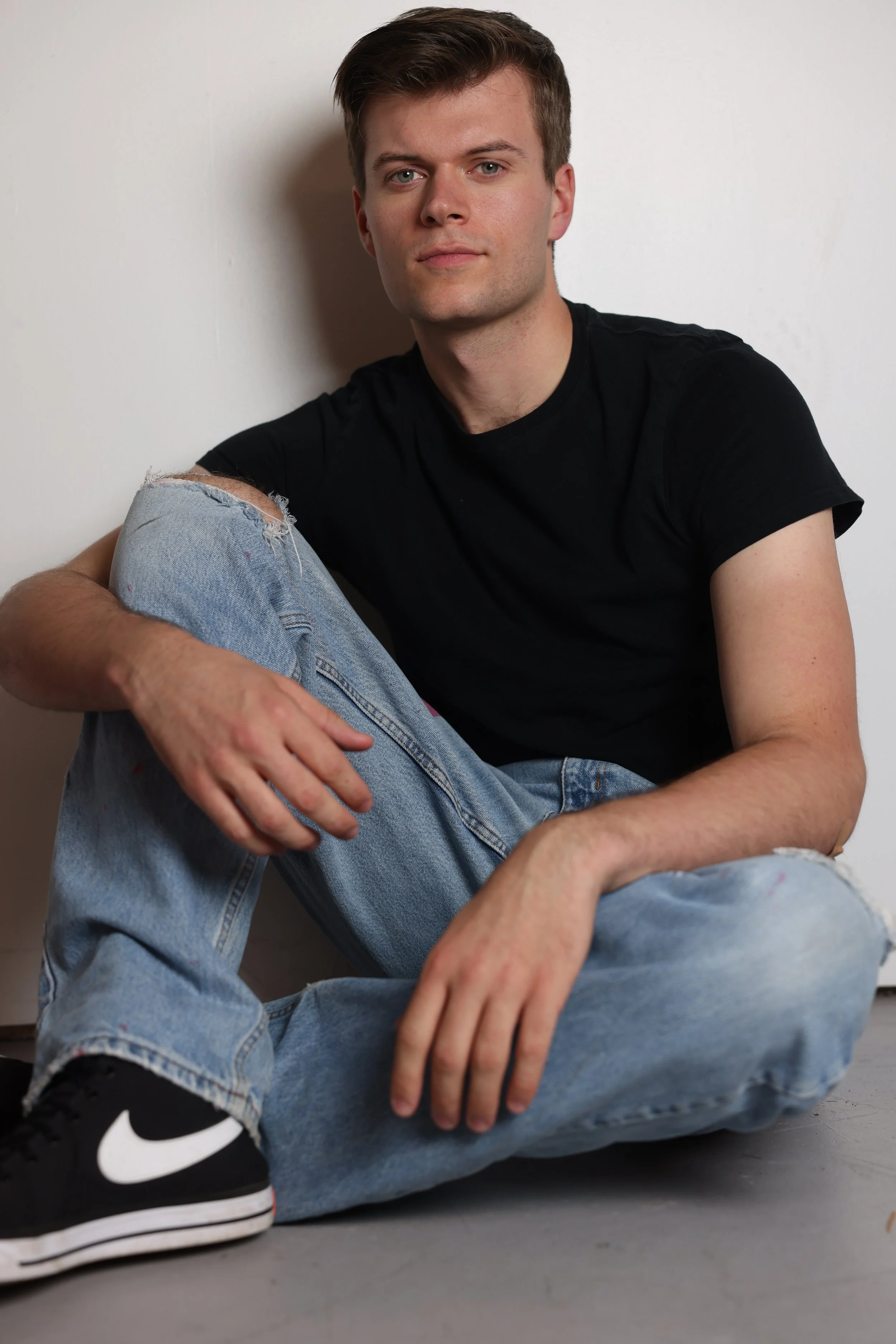 A young man sitting on the floor against a white wall, wearing a black t-shirt, ripped jeans, and Nike sneakers, looking at the camera.