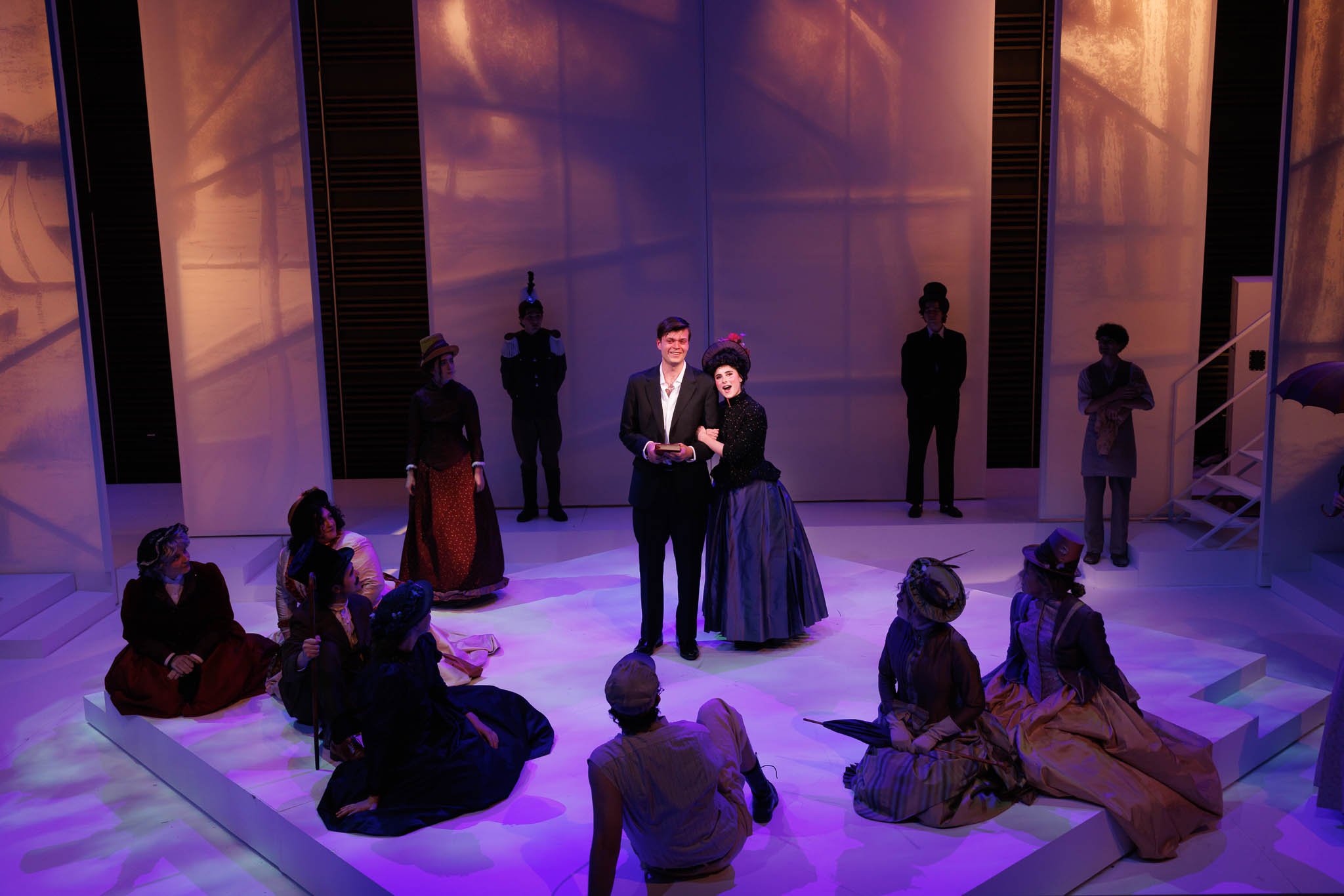 Theatrical performance with actors on stage; a man and a woman in vintage costumes singing or speaking, surrounded by other actors seated and standing, with stage lighting and set design in the background.