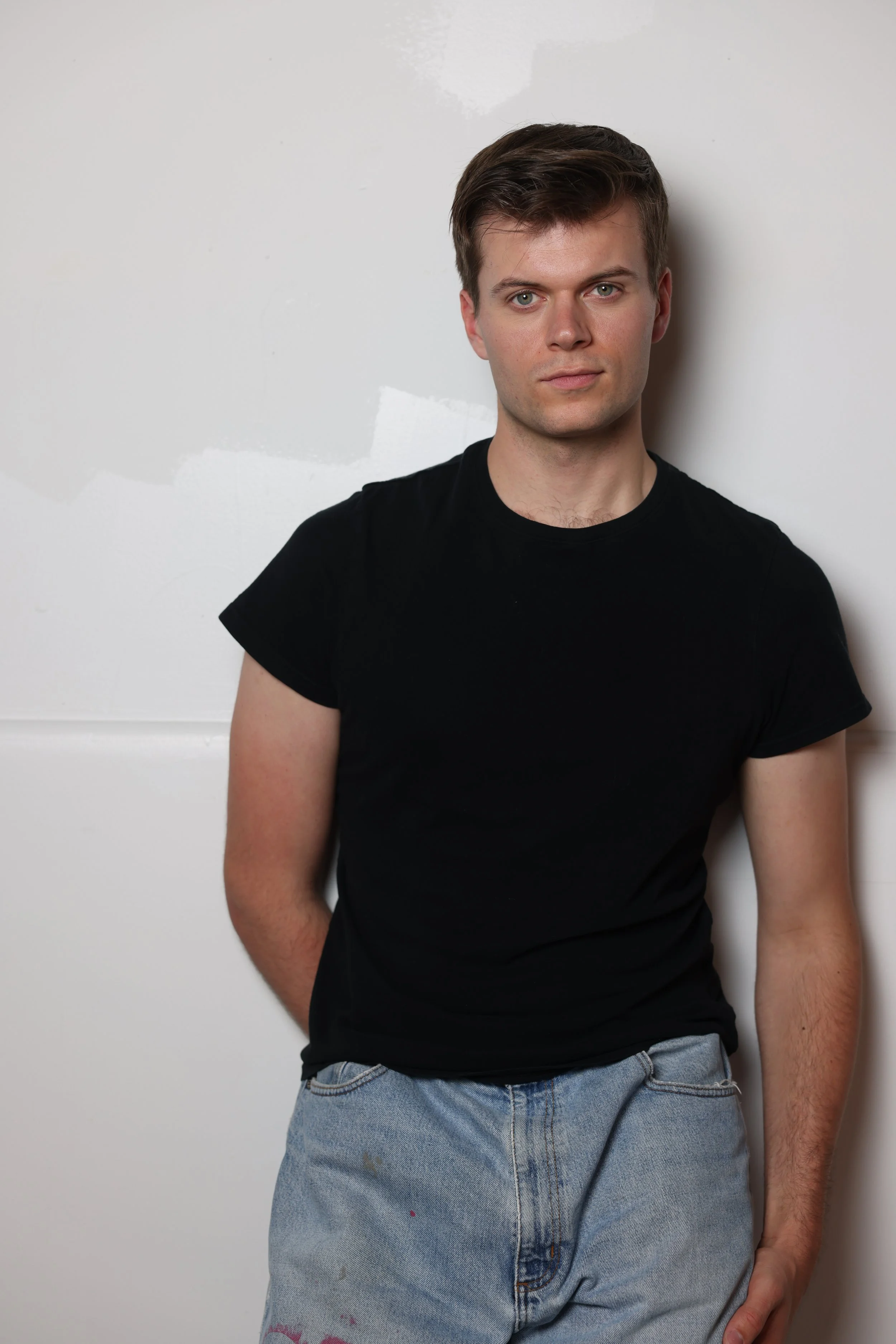 A young man with short brown hair, wearing a black T-shirt and light blue jeans, leaning against a white wall.