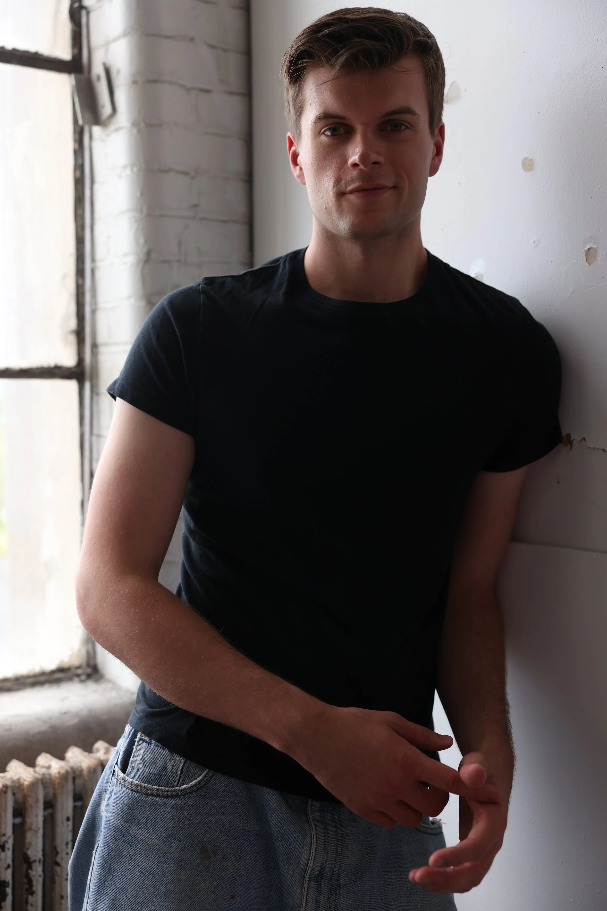 A young man with light brown hair, wearing a black t-shirt and light blue jeans, standing indoors near a white wall and a window with blurred outdoor scenery.