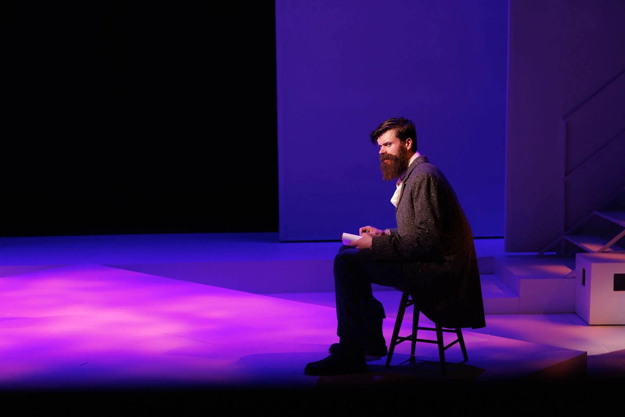 A man with a beard wearing a gray coat and white shirt sitting on a stool on a stage with purple lighting.