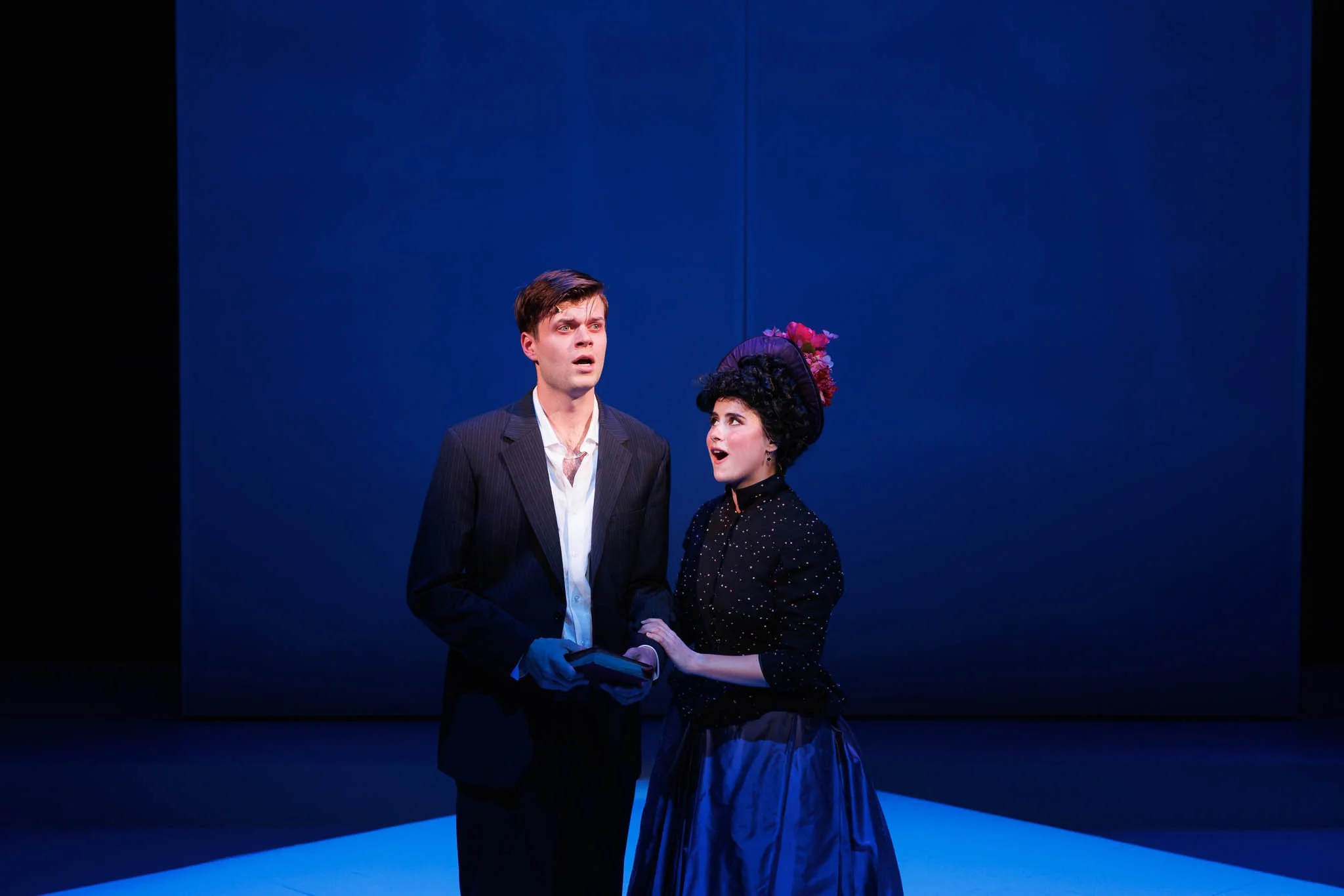 Two actors on stage, one man in a black suit holding a book and a woman in a black dress with a purple hat, singing or speaking with emotional expressions, against a solid blue background.