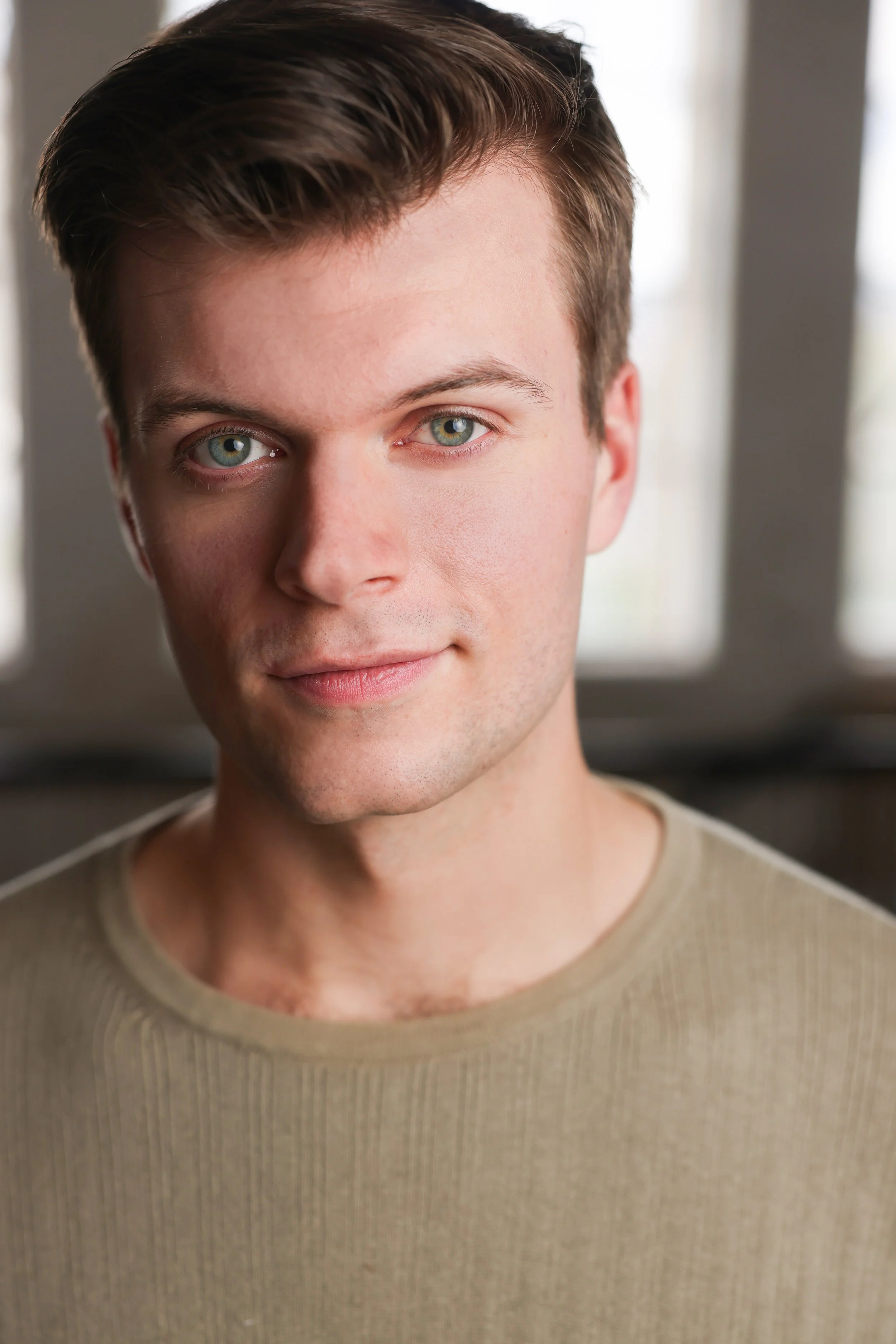 Close-up of a young man with fair skin, blue-green eyes, brown hair, wearing a beige shirt, indoors with natural light.