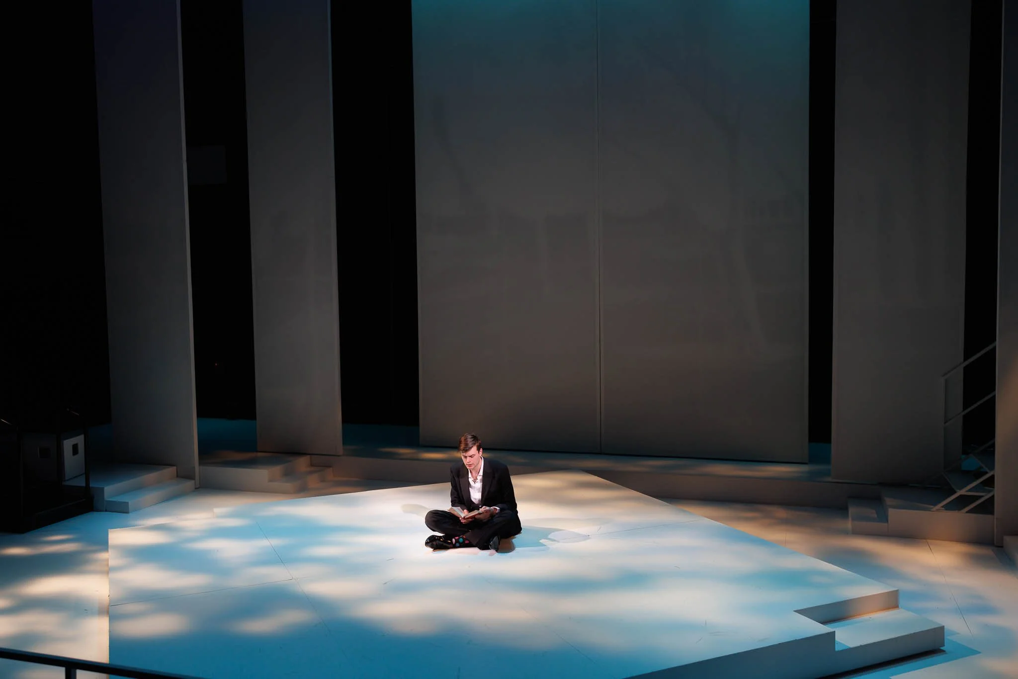 A person sitting cross-legged on a stage reading a book, with a modern minimalist set design and soft lighting.