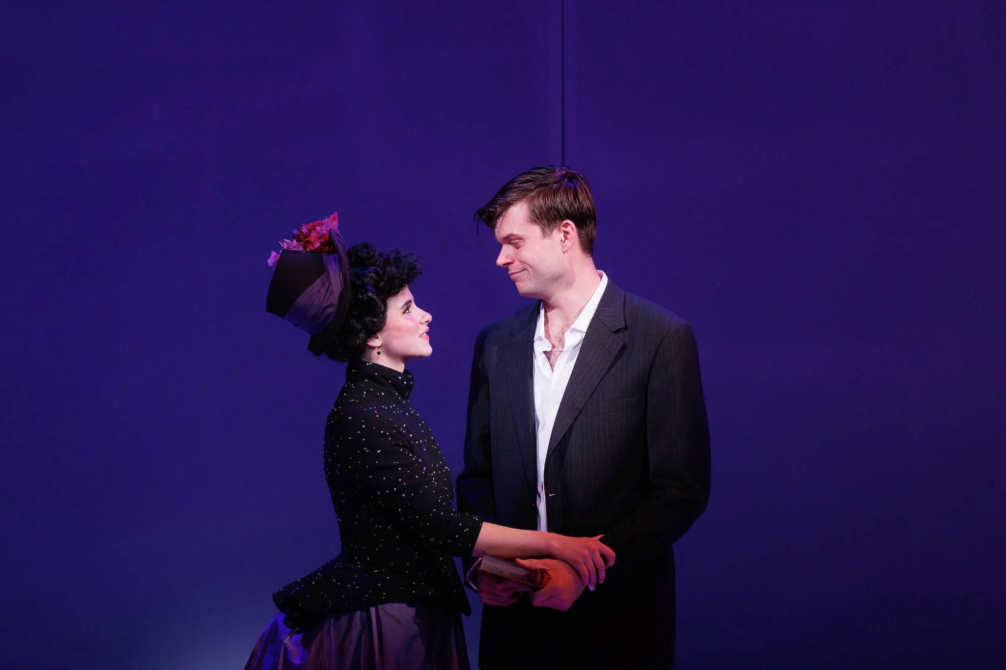 A man and woman stand facing each other, holding hands, against a plain purple background. The woman wears a black dress with sparkles and a large purple hat with flowers. The man wears a dark suit and white shirt, looking at her with a smile.