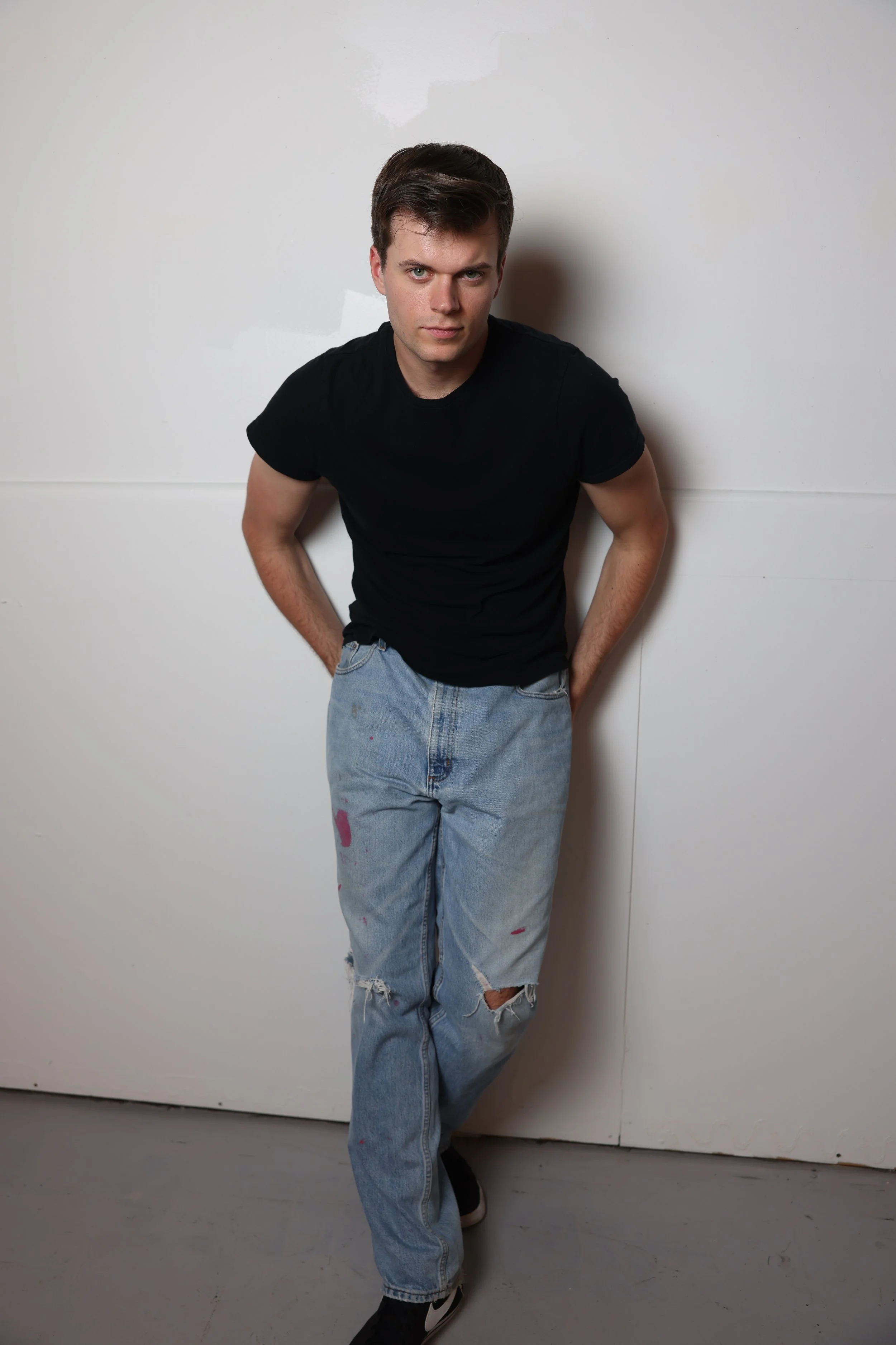 A young man with dark hair wearing a black T-shirt and distressed light blue jeans standing against a plain white wall.