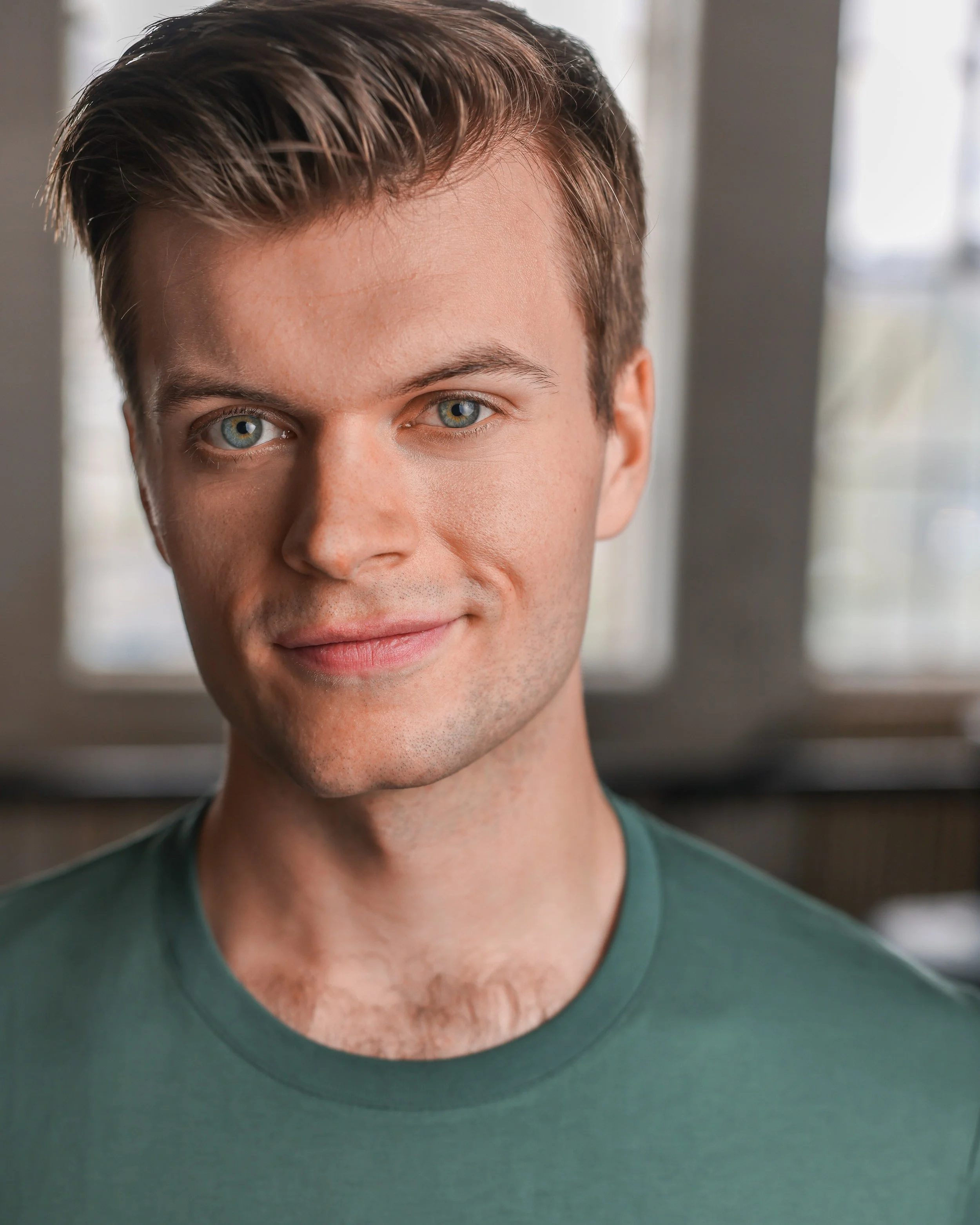 Close-up portrait of a young man with blue eyes and light brown hair smiling, wearing a green t-shirt, in front of a window with natural light.