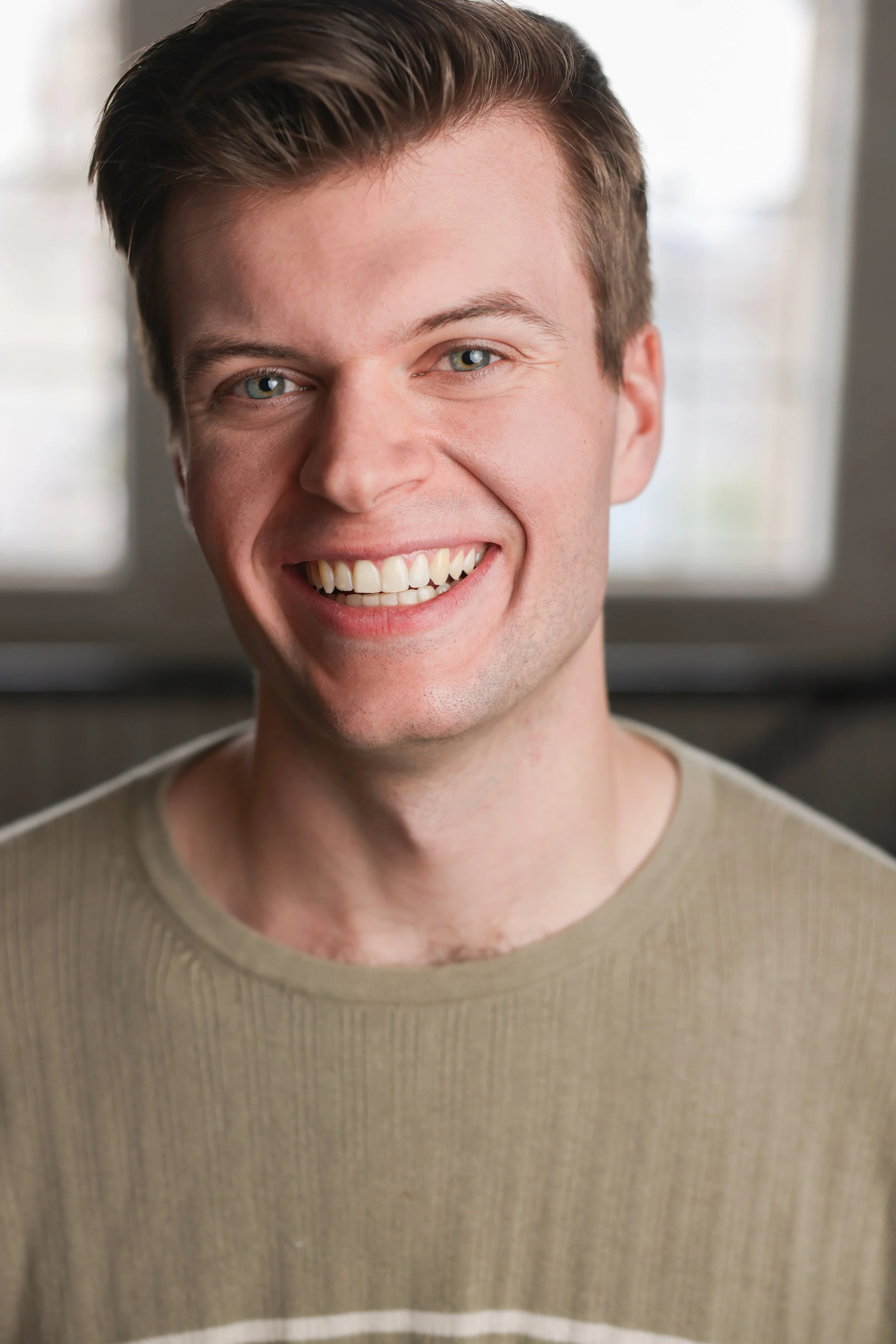 A young man with short brown hair and blue eyes smiling at the camera, wearing a beige sweater, standing indoors near a window.