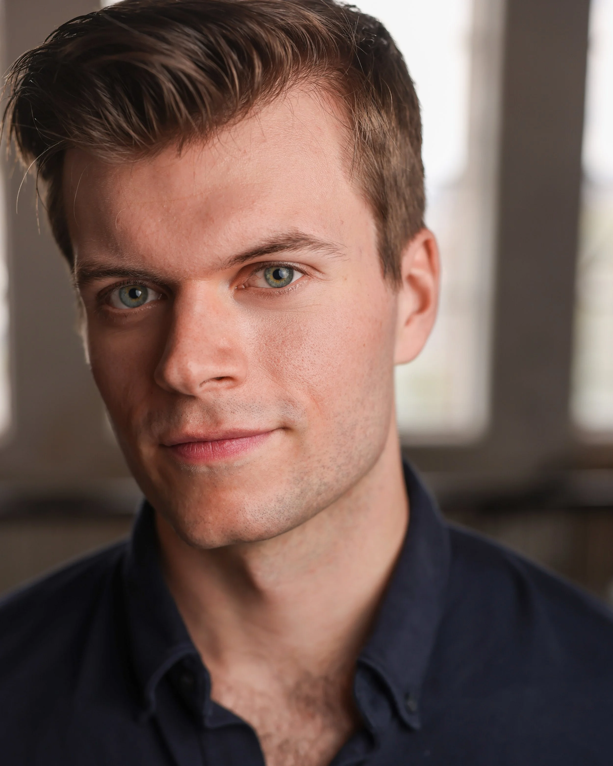 Close-up portrait of a young man with short brown hair, blue eyes, and light skin, wearing a dark shirt, inside a room with windows in the background.