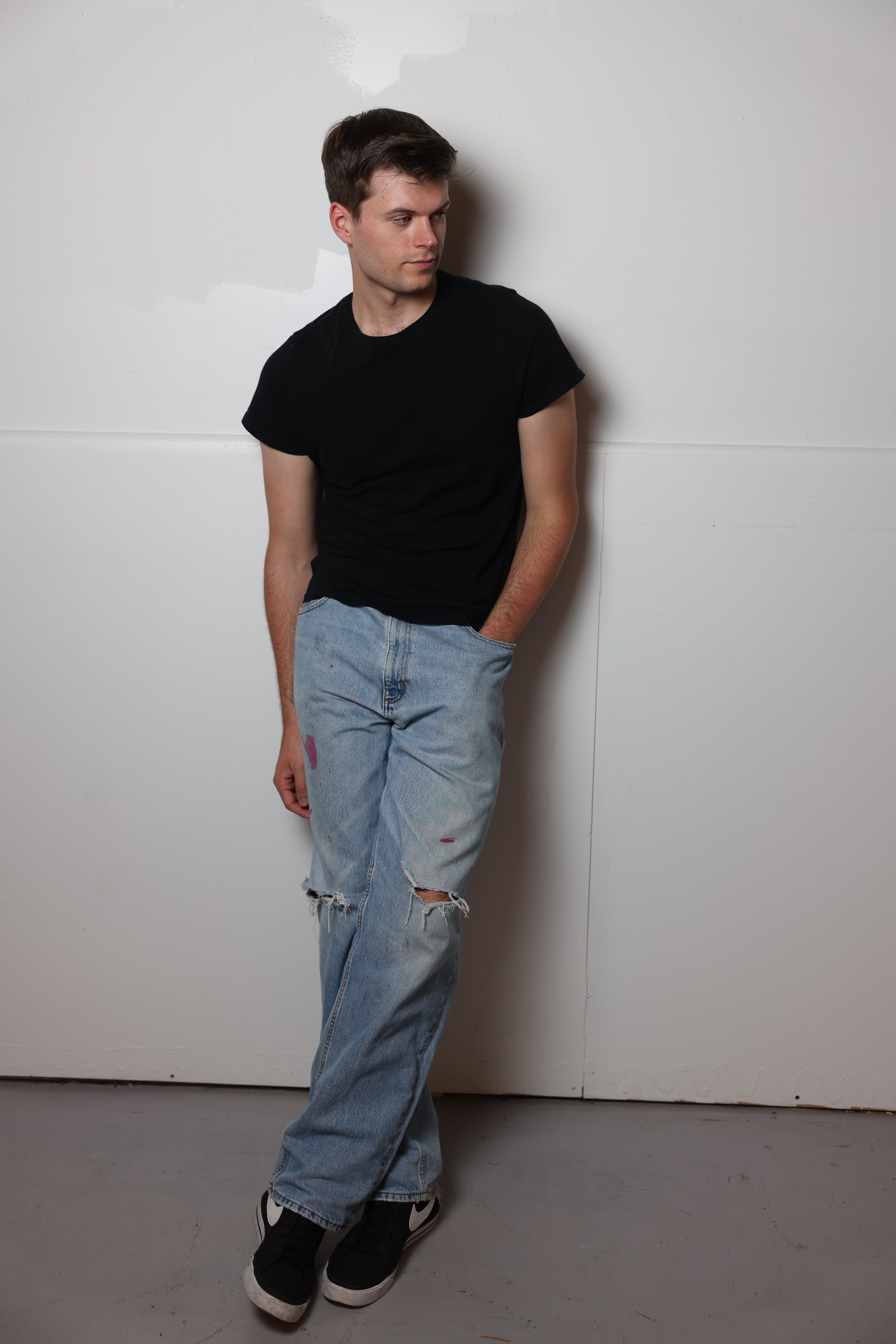 Young man with short brown hair wearing a black T-shirt, ripped blue jeans, and black Nike sneakers standing against a white wall with his left hand in his pocket and looking to the side.