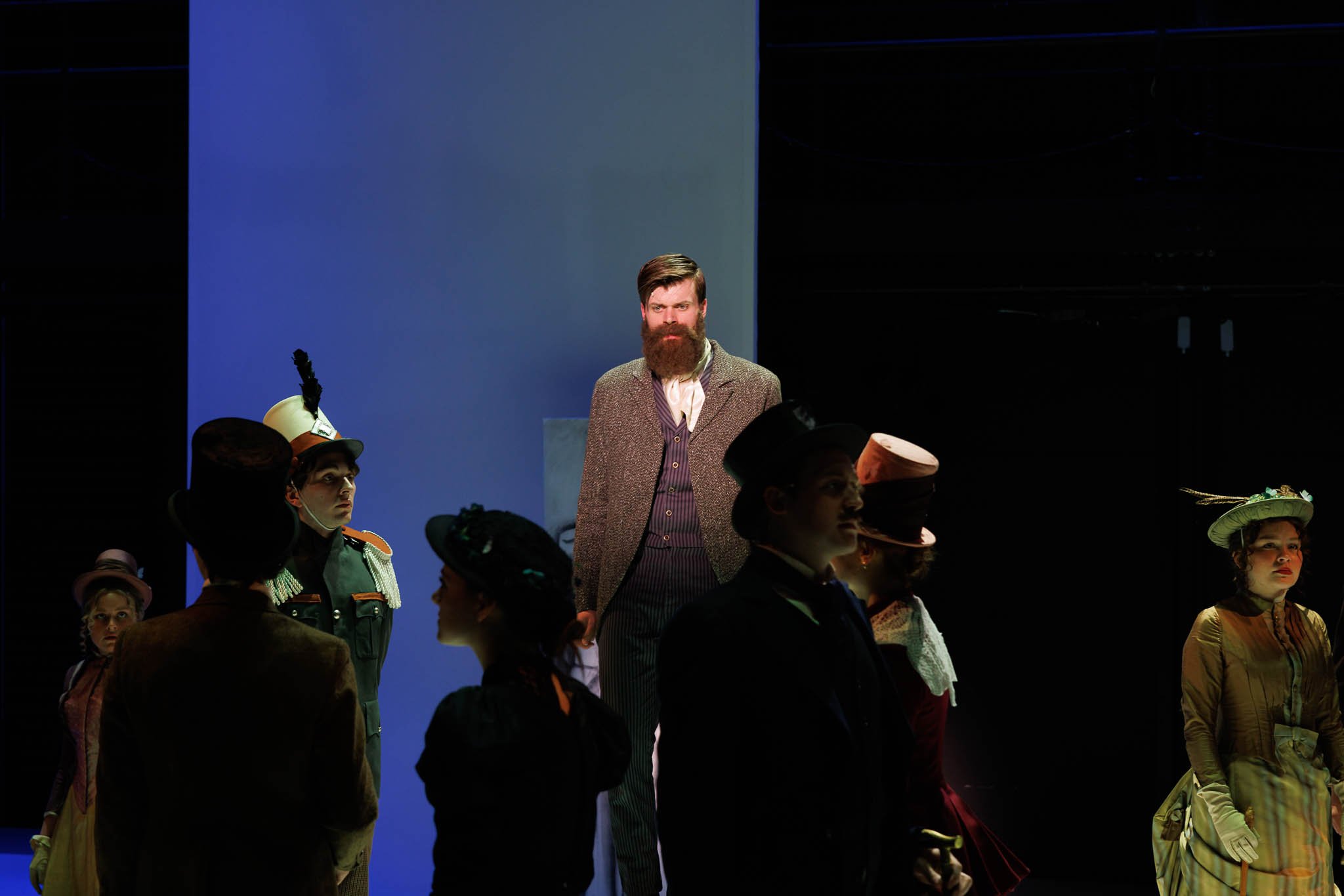 Actors performing on stage in period costumes with a blue and black background, featuring a man with a beard in the center and women in historical attire around him.