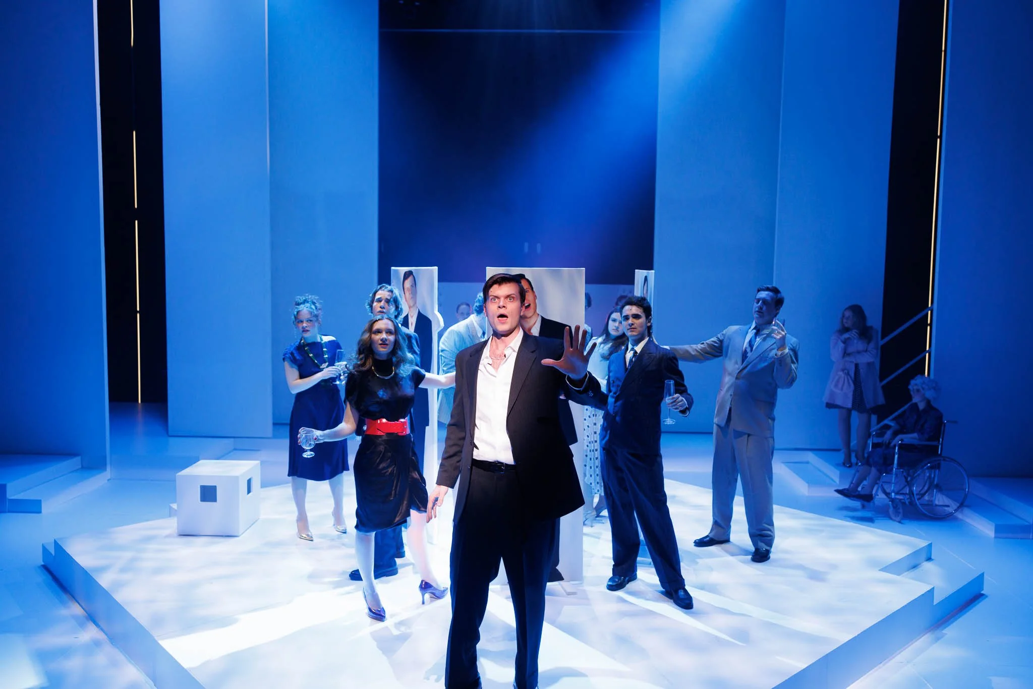 A theatrical stage with actors in formal attire, some holding glasses, performing a scene. The stage has a minimalist, modern design with blue lighting and a white floor.