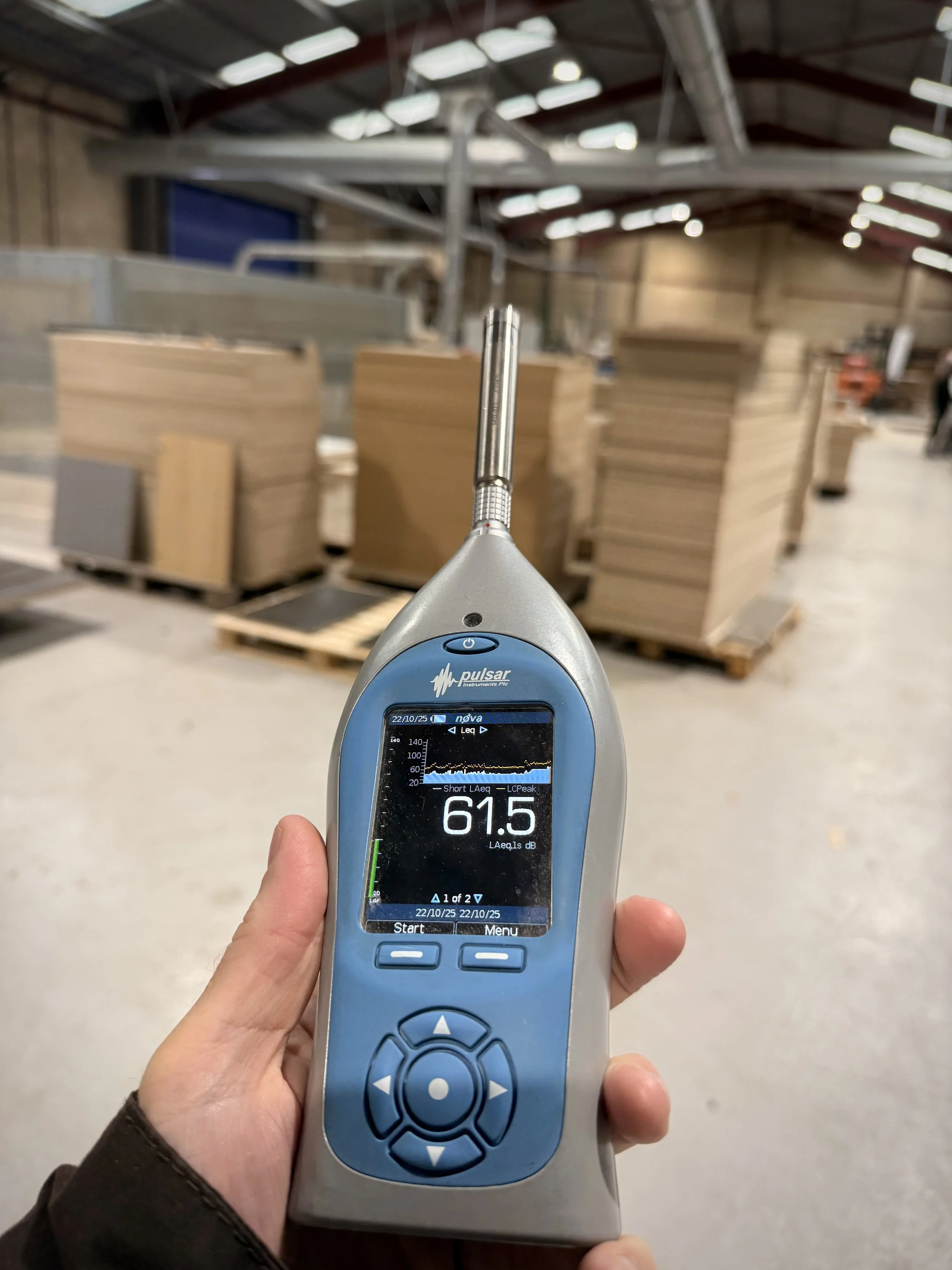 Noise meter being held in the hand in a noise assessment for a joinery workshop, with large sheets of wood behind