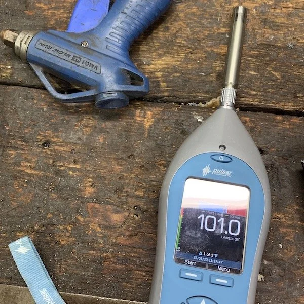 Noise meter reading a high noise level of 101 dB(A) on a noise assessment, on a wooden bench next to an air gun with an air leak