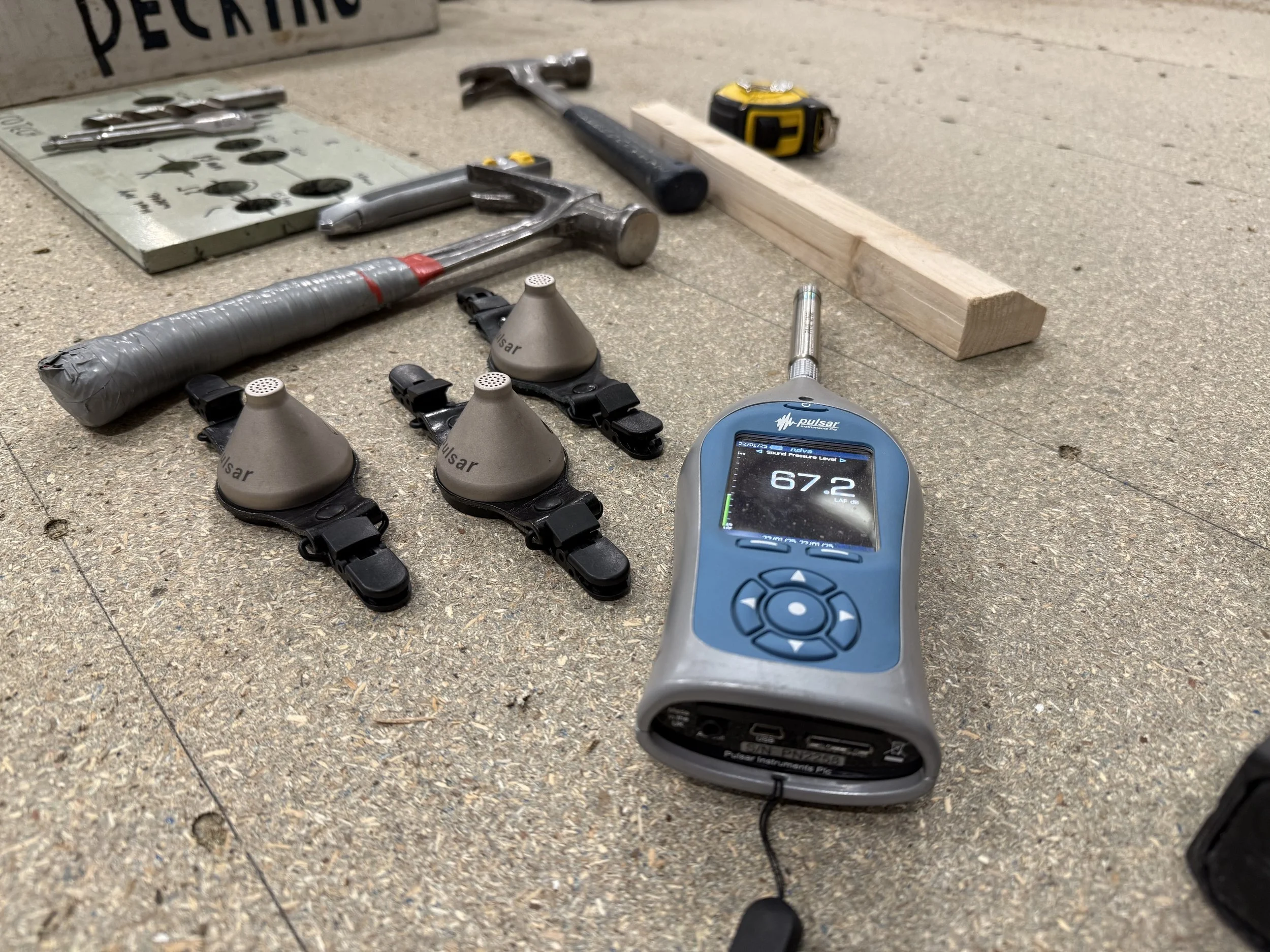 A noise meter and three dosemeters on a table next to hammers in a joinery workshop noise assessment