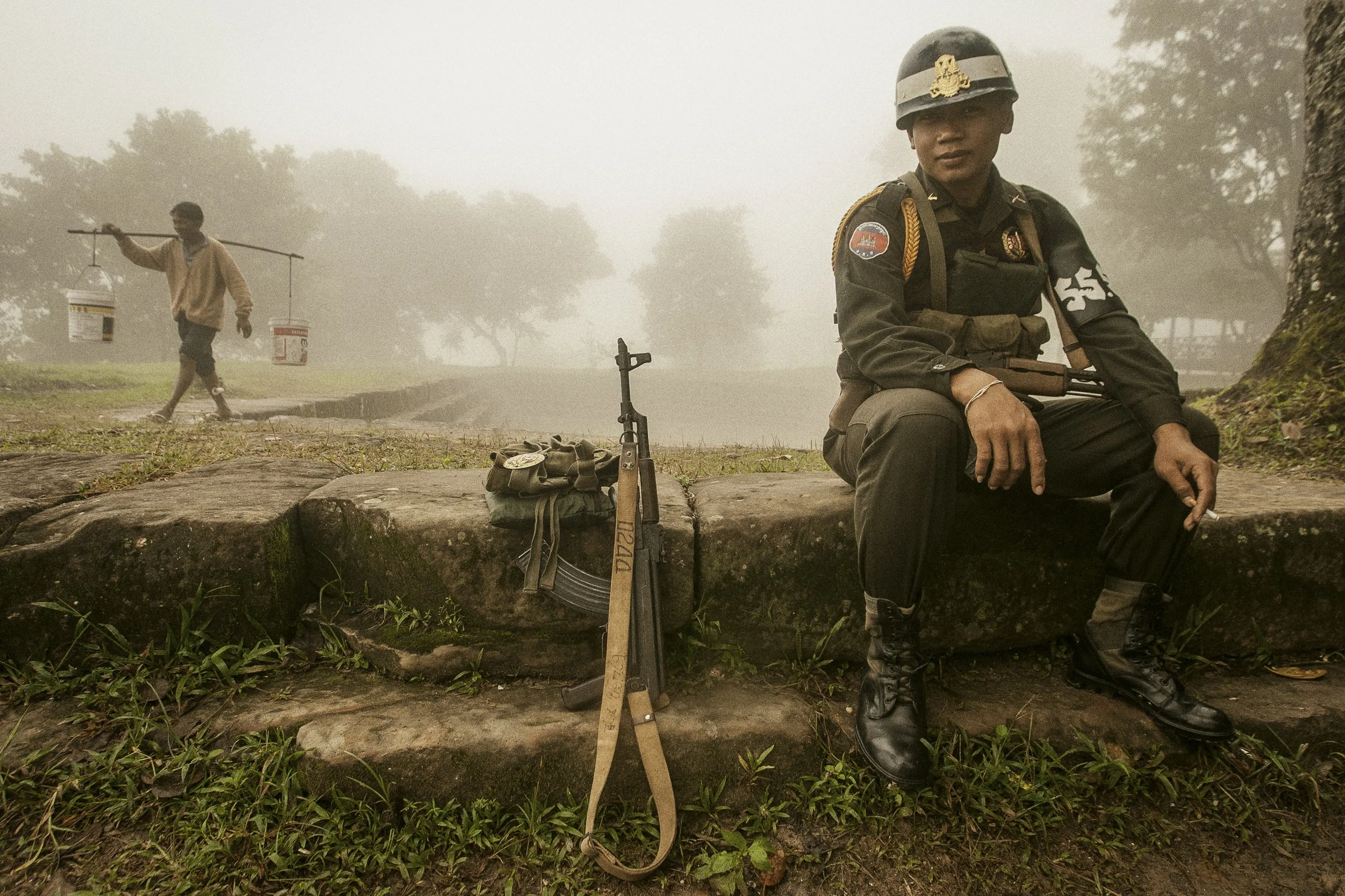 An RCAF Military Policeman enjoys a morning cigarette as a local villager collects water from the nearby Trapeang, an ancient small water reservoir.