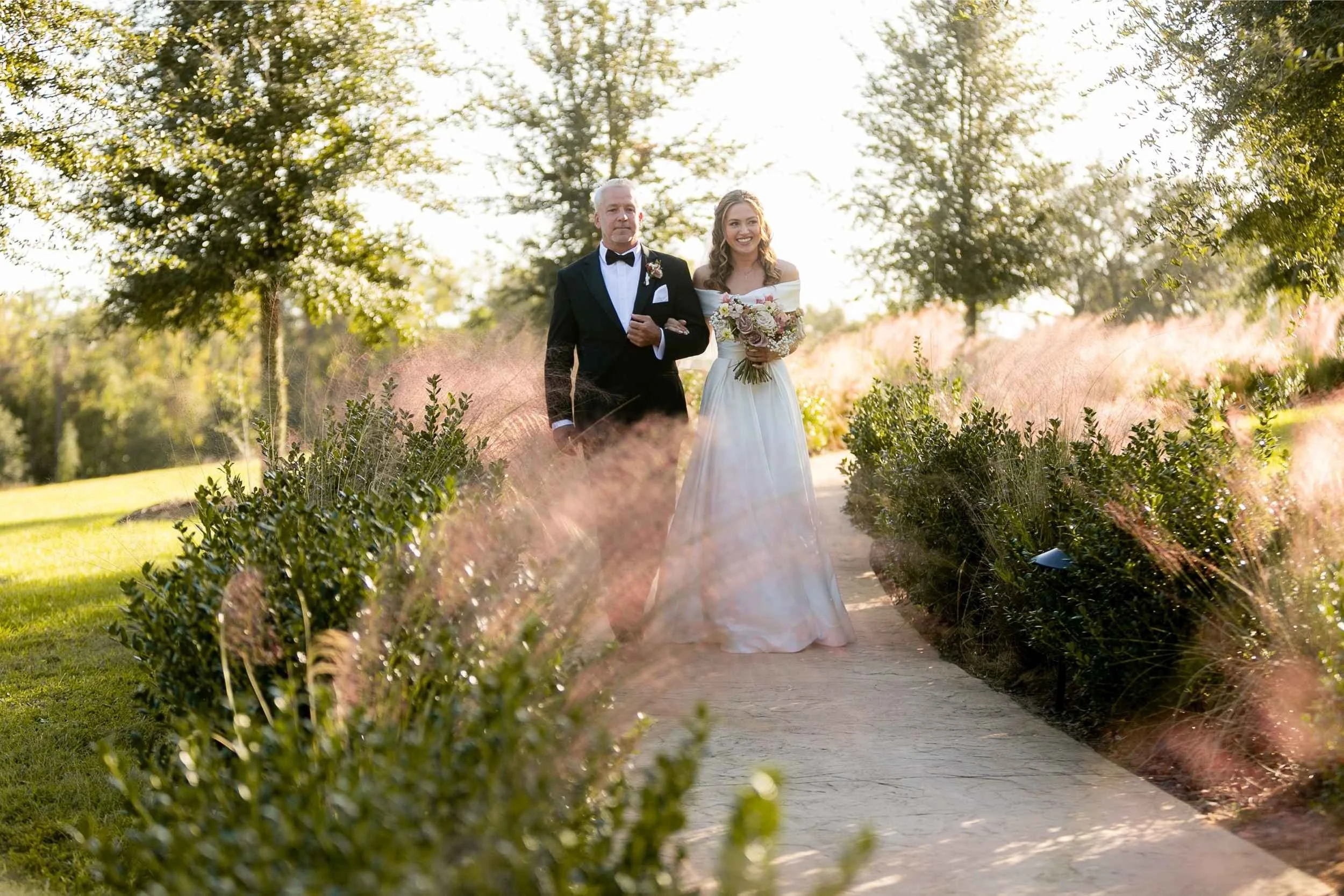 A bride in a white dress holding a bouquet walking down a garden path with a man in a tuxedo during sunset for a wedding photographed by Cannonfire Photography.