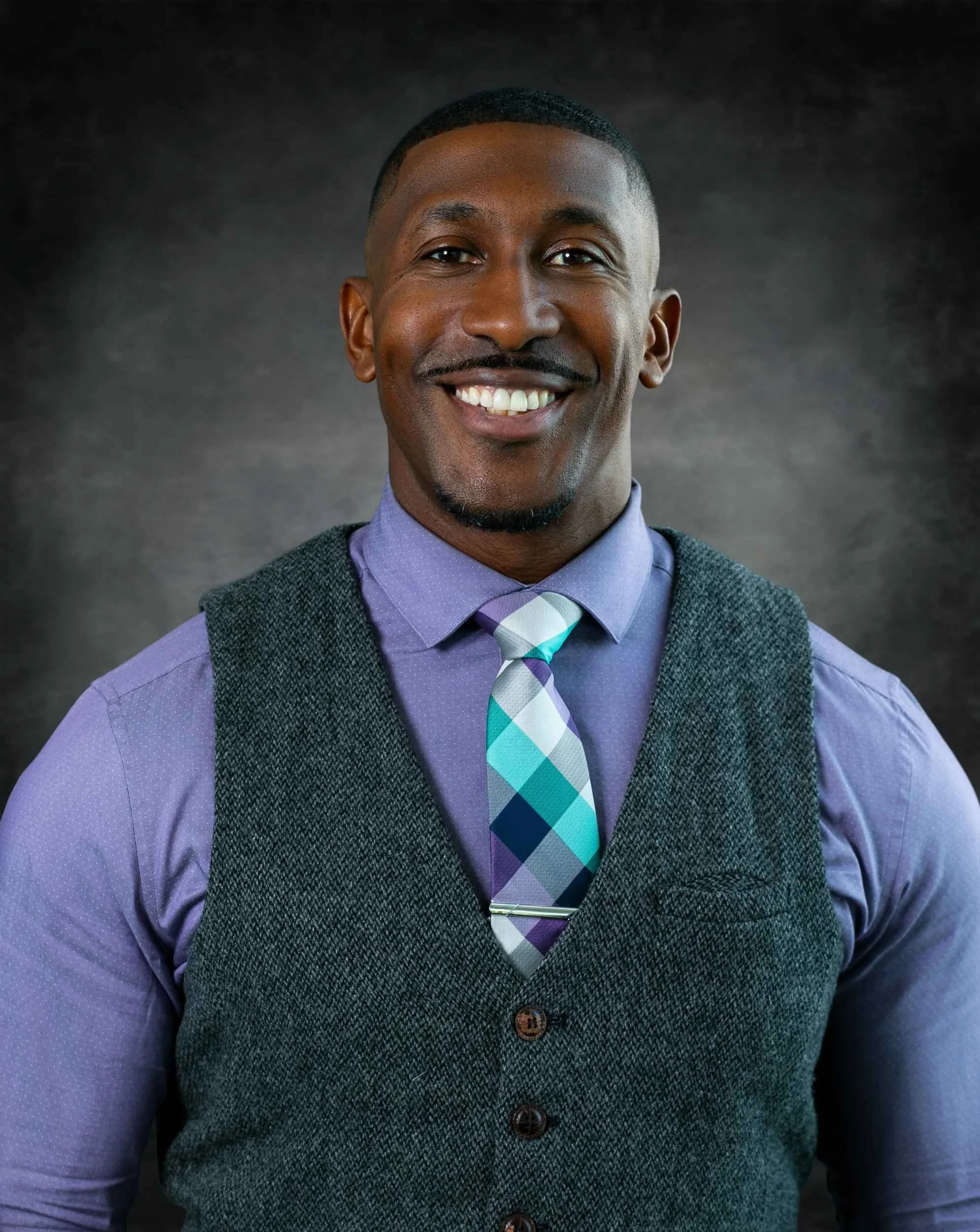 Portrait of a smiling Black man wearing a purple dress shirt, patterned tie, and gray vest, against a dark background for a headshot.