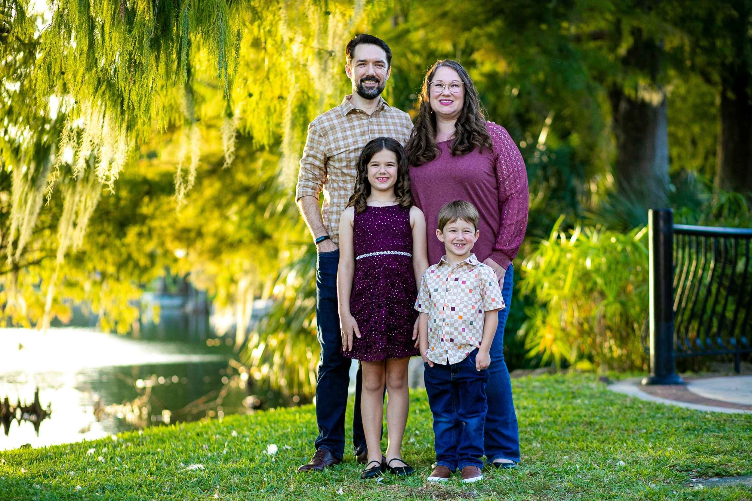 A family of four standing on grass near a body of water with trees in the background, smiling outdoors on a sunny day during a family portrait session with Cannonfire Photography.