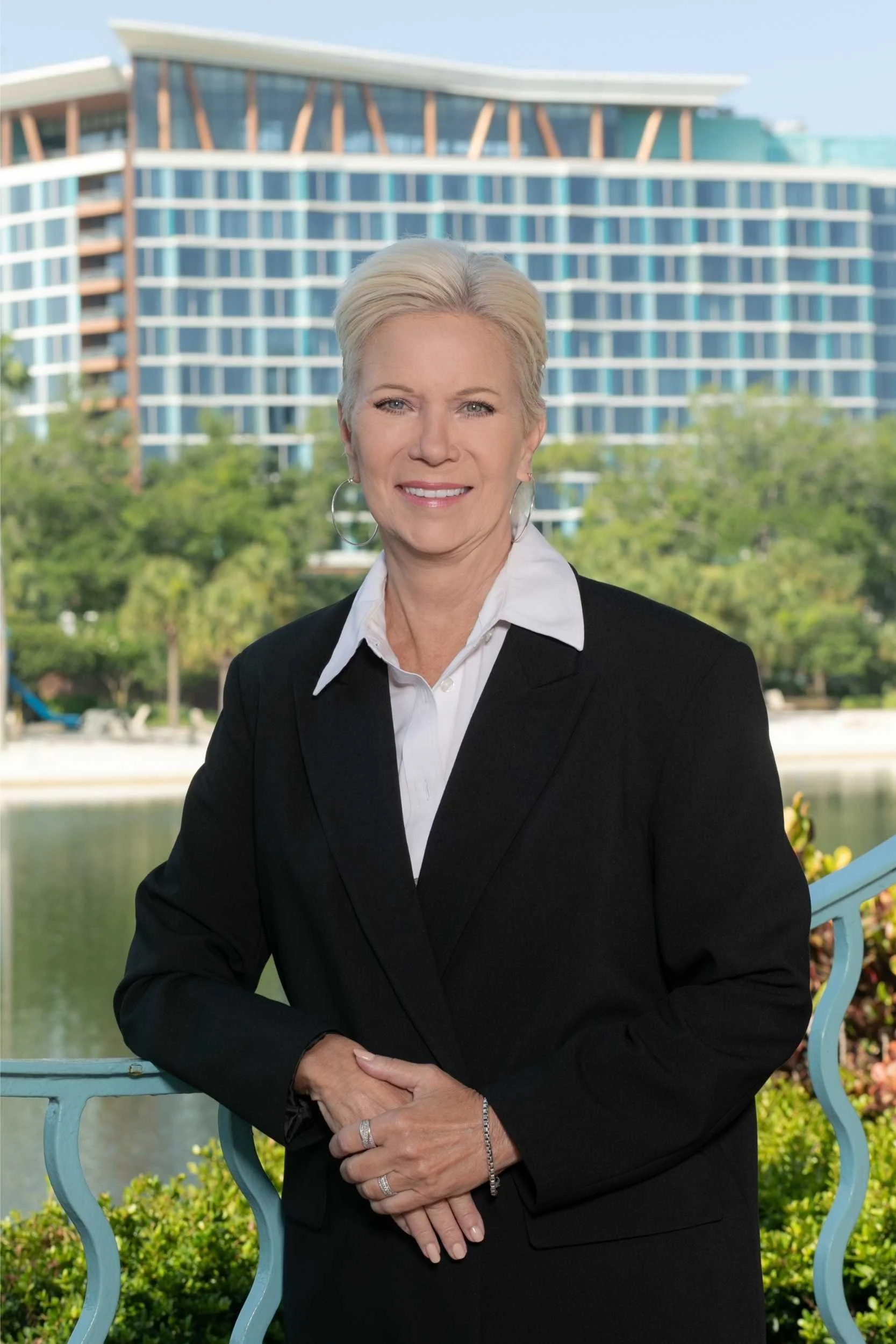 A woman with short blonde hair smiling and wearing a black blazer over a white shirt, standing outdoors in front of a modern building with glass windows and a body of water with trees in the background for a headshot with Cannonfire Photography.