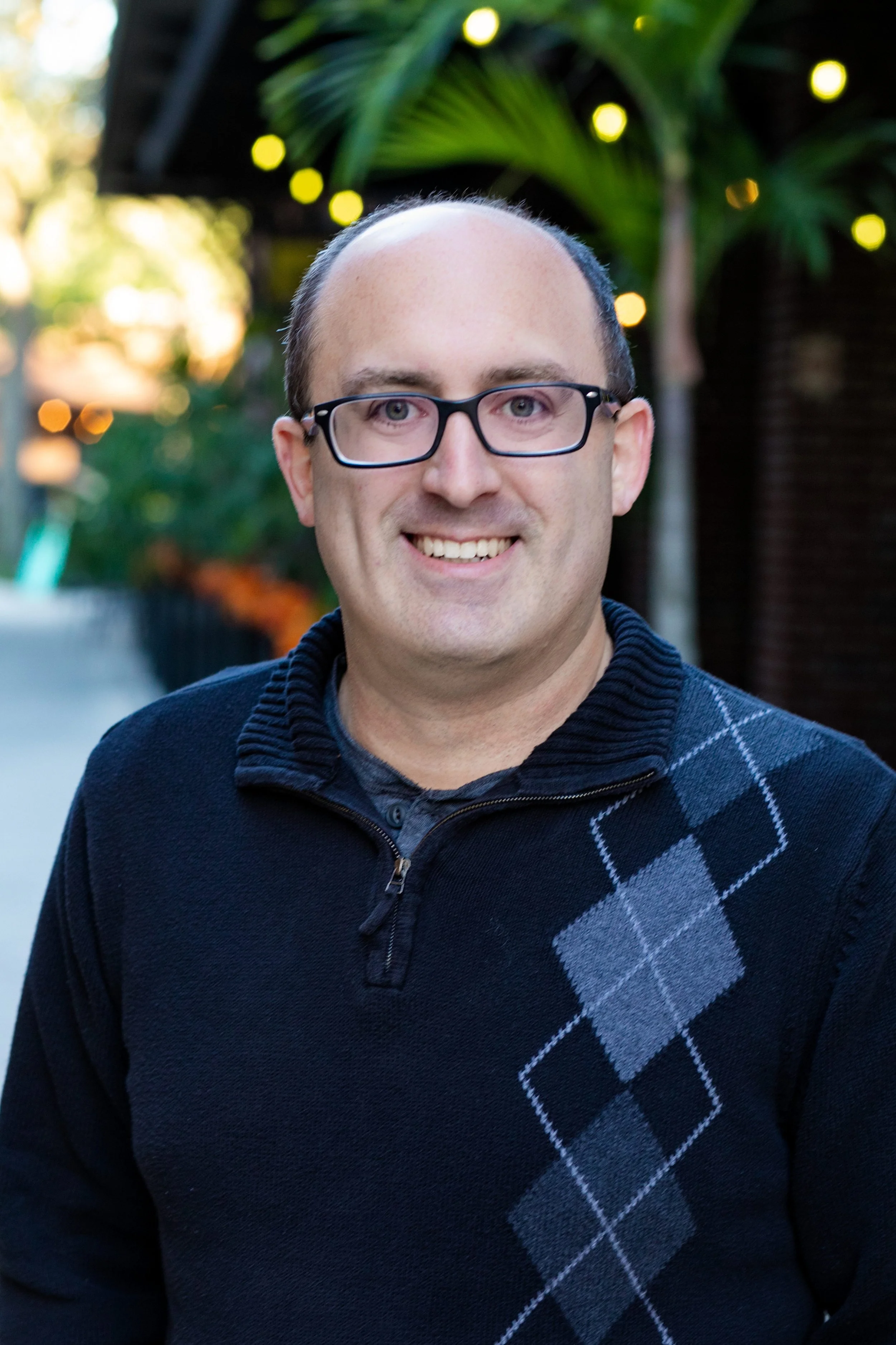 A smiling man with glasses and a dark sweater standing outdoors with blurred greenery and lights in the background for a headshot with Cannonfire Photography in the West Orange area of Central Florida.