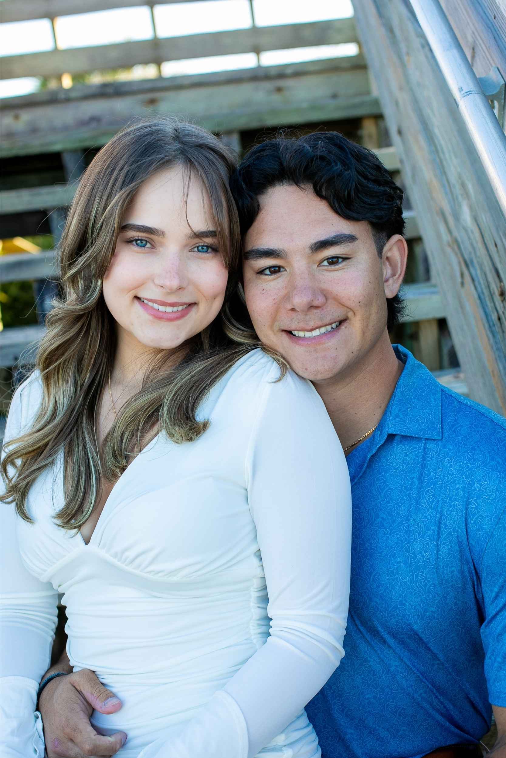 A young woman with long, wavy light brown hair and blue eyes, smiling, is sitting close to a young man with short, black wavy hair and brown eyes, also smiling. They are outdoors near wooden stairs.