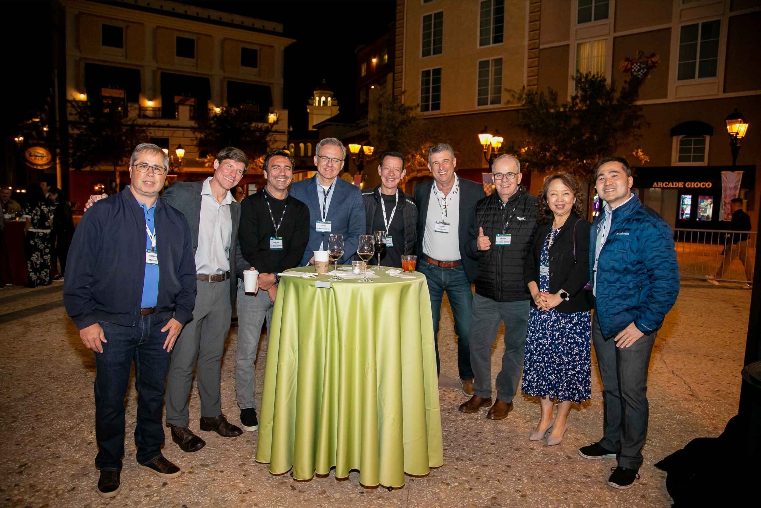 Group of ten people gathered around a table outdoors at night, smiling, with drinks, in a festive setting with buildings and lights in the background.