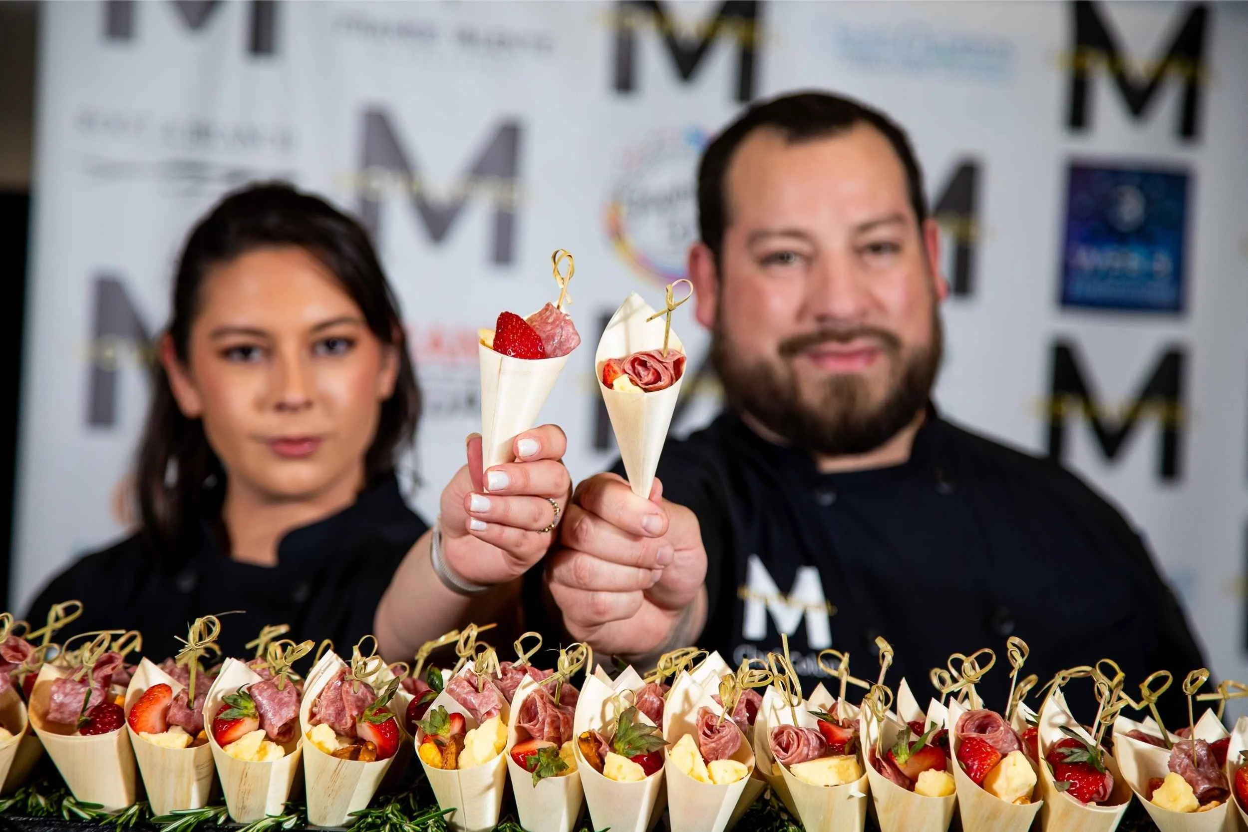 Two individuals holding small food cups with various appetizers, standing behind a table filled with similar appetizers.