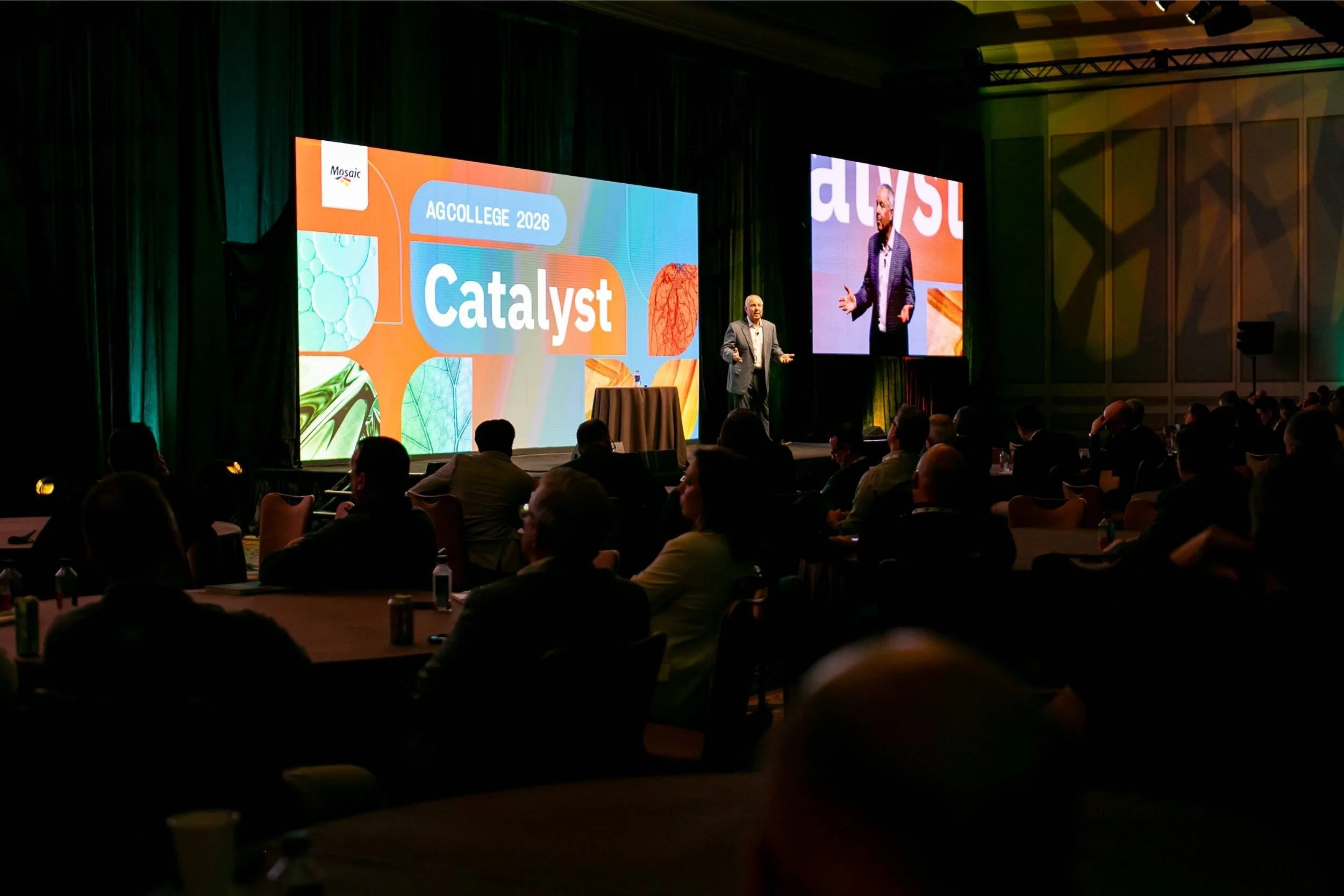 A man giving a presentation on a stage at a conference, with a large screen displaying 'AGCOLLEGE 2026 Catalyst' and a smaller screen showing the presenter, in a dimly lit conference room filled with seated attendees.