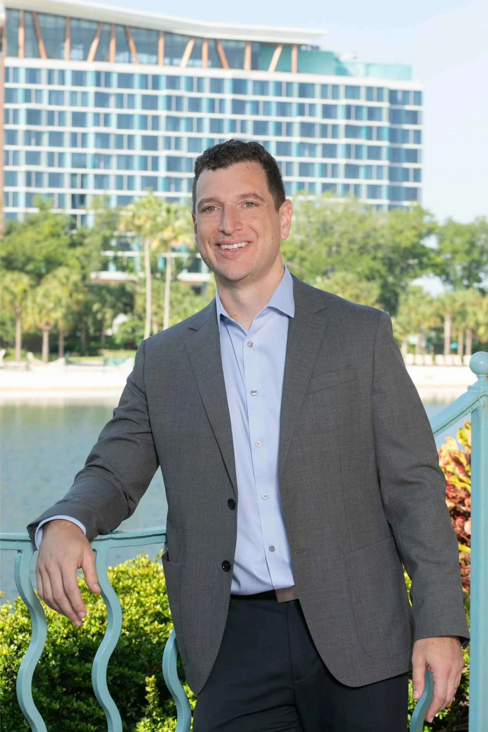 A man in a gray suit and light blue shirt standing outdoors by a teal-colored railing, with a cityscape and trees in the background for a headshot with Cannonfire Photography.