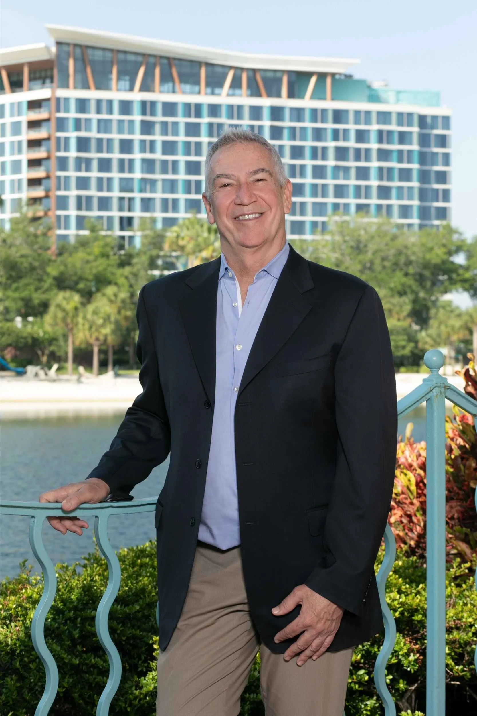 A smiling man in a black blazer and light blue shirt standing outdoors near water, with green trees and a modern building in the background for a headshot with Cannonfire Photography.