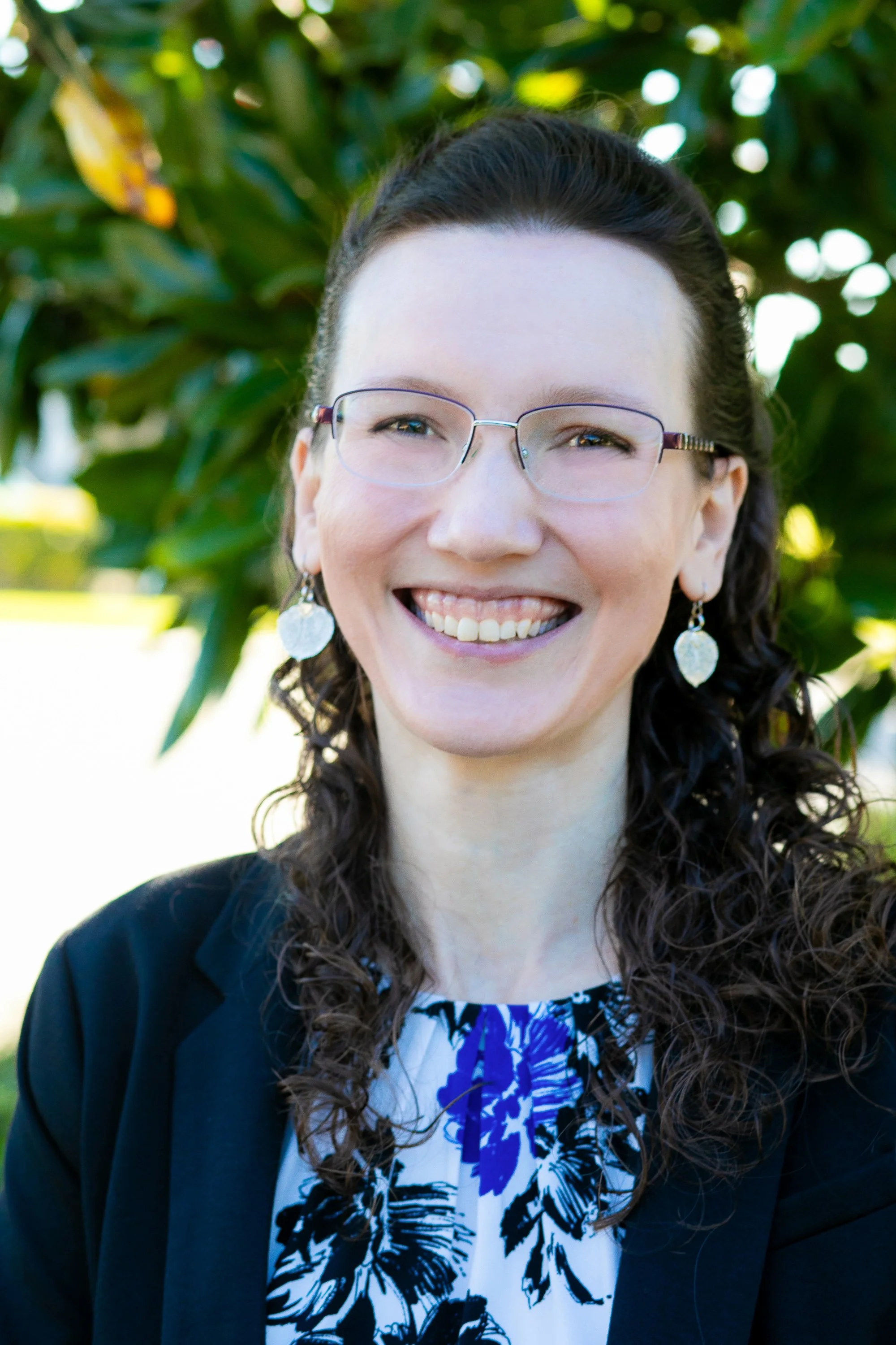 A woman with glasses and earrings smiling outdoors with greenery in the background for a headshot with Cannonfire Photography in Orlando.