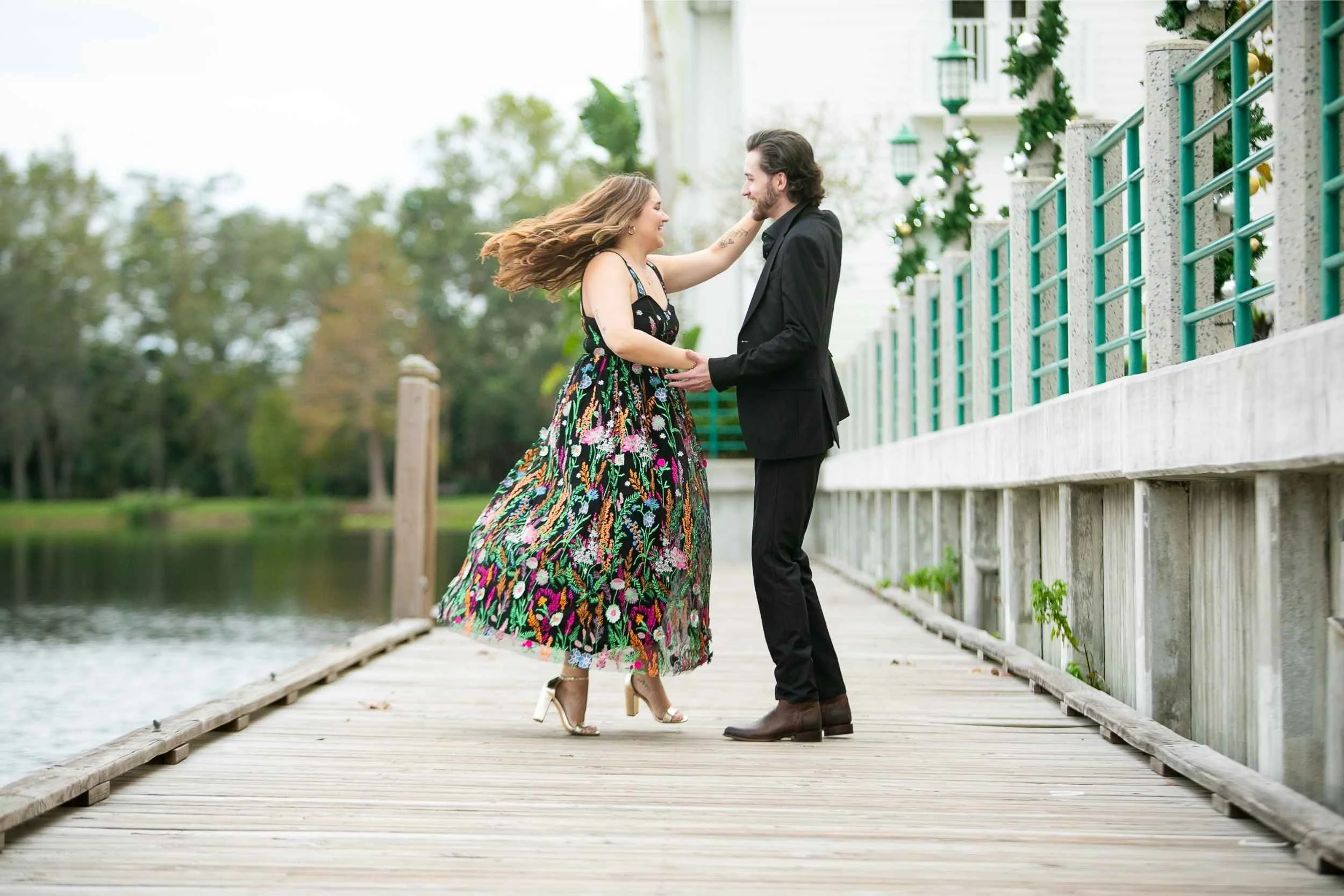 A couple is dancing and smiling on a wooden bridge beside a lake, surrounded by trees and a white house with green railings in the background for an engagement photography session with Cannonfire Photography.