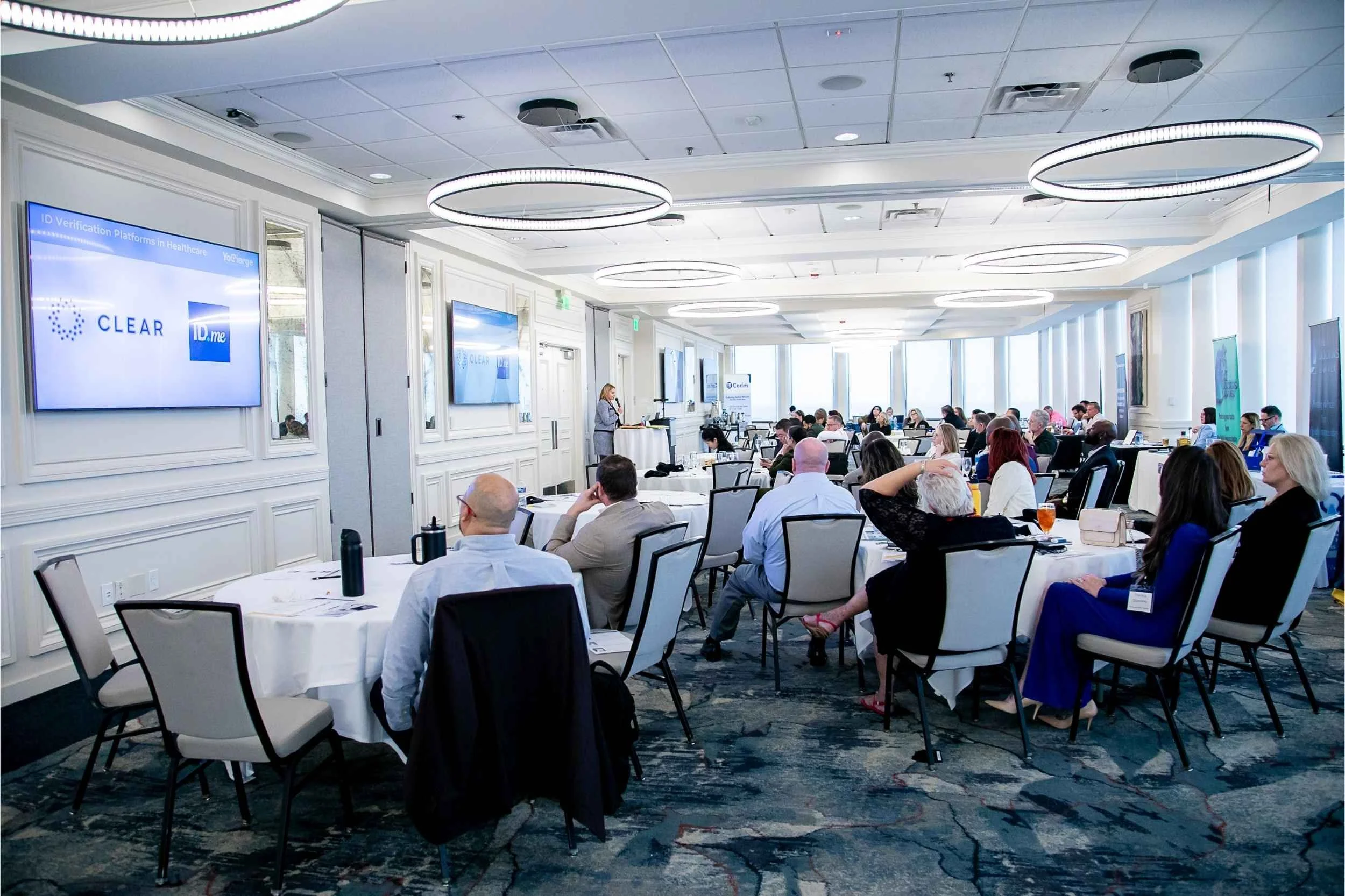 A conference room filled with seated attendees, facing a presenter at a podium. Multiple screens display a slide with the logos 'CLEAR' and 'ID.me'. Bright, modern interior with overhead lighting and large windows.
