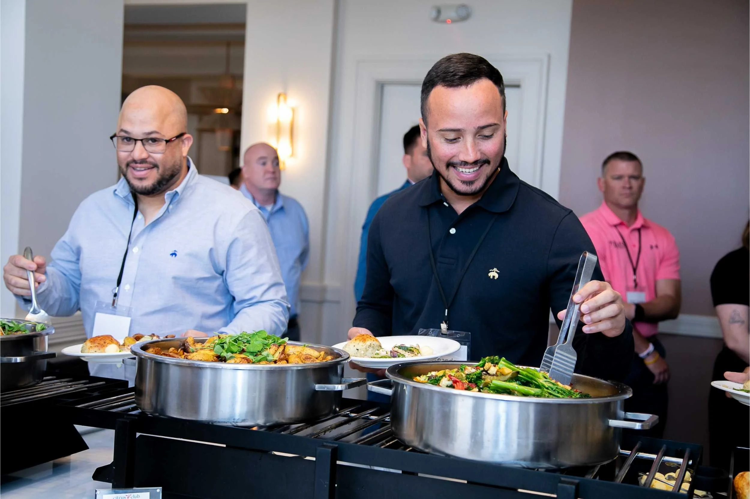 Two men serving themselves from buffet dishes at a social event, with other guests in the background for a conference photographed by Cannonfire Photography in Orlando.