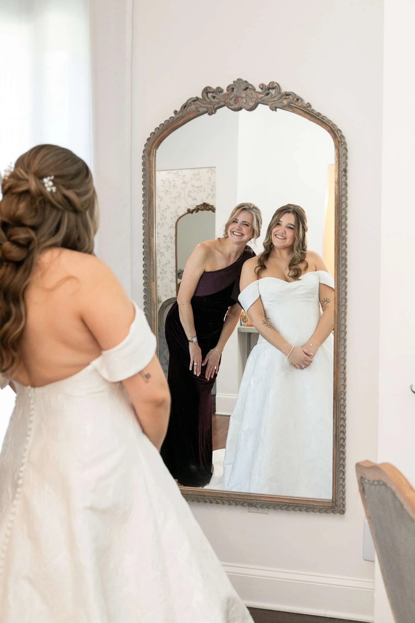 A young woman in a white off-shoulder wedding dress looks at herself in a mirror, with her mother in a dark purple dress smiling behind her for a wedding photographed by Cannonfire Photography.