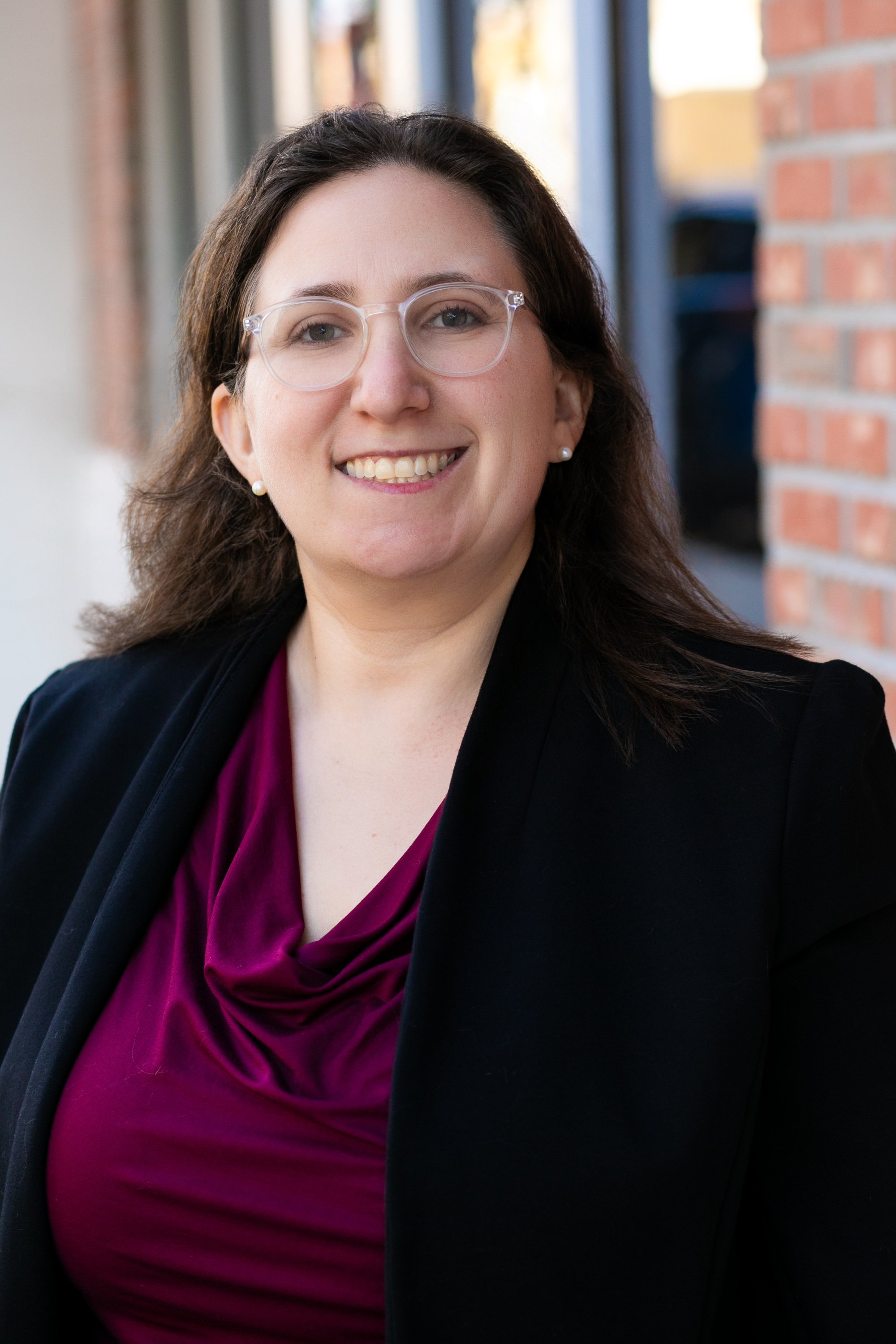A woman with brown hair, glasses, and a bright smile wearing a black blazer and purple blouse, standing outdoors near a brick building for a headshot with Cannonfire Photography in the West Orange area of Central Florida.
