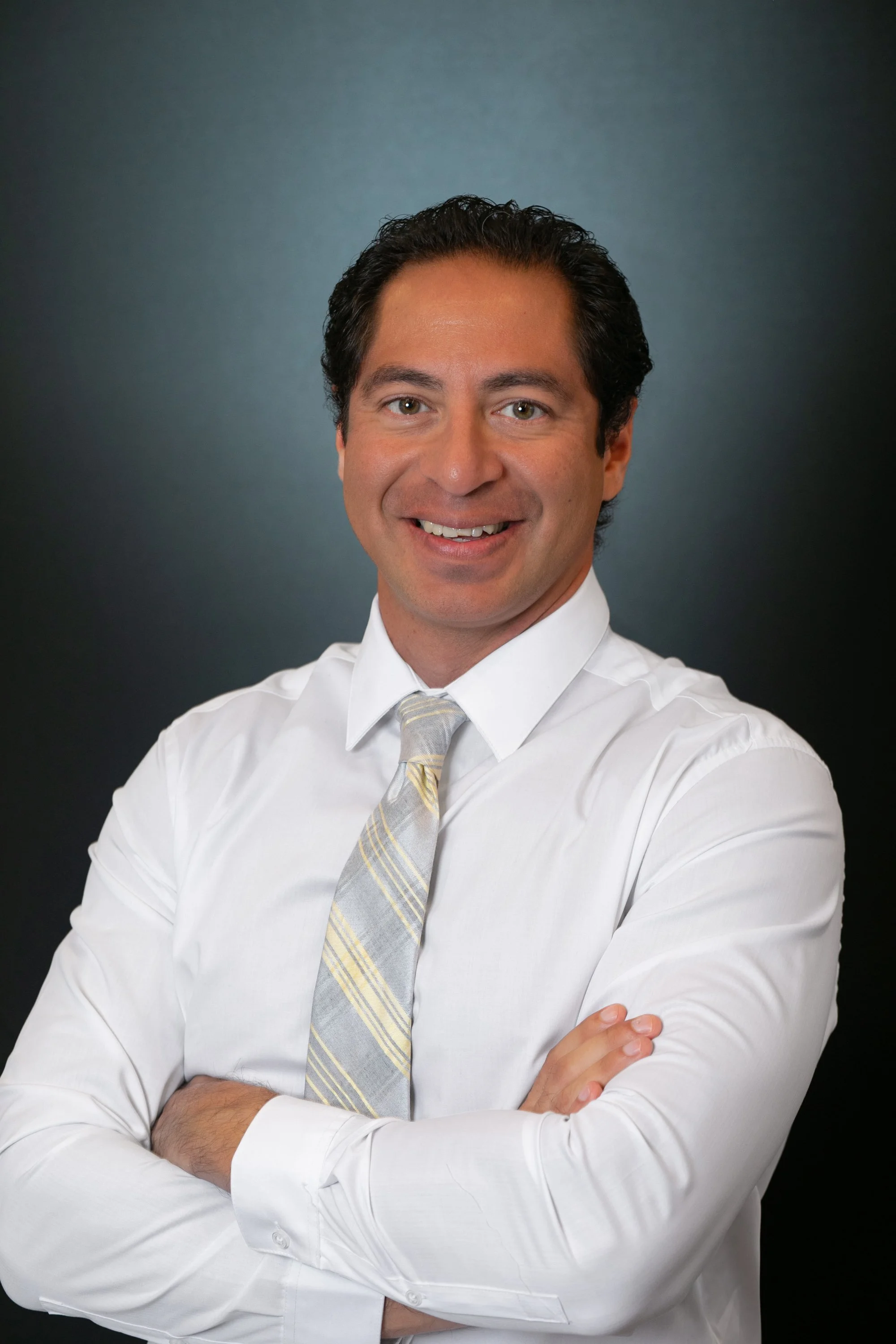 A smiling man with dark hair in a white shirt and patterned tie, standing with his arms crossed against a dark gradient background for a headshot with Cannonfire Photography in Orlando;.
