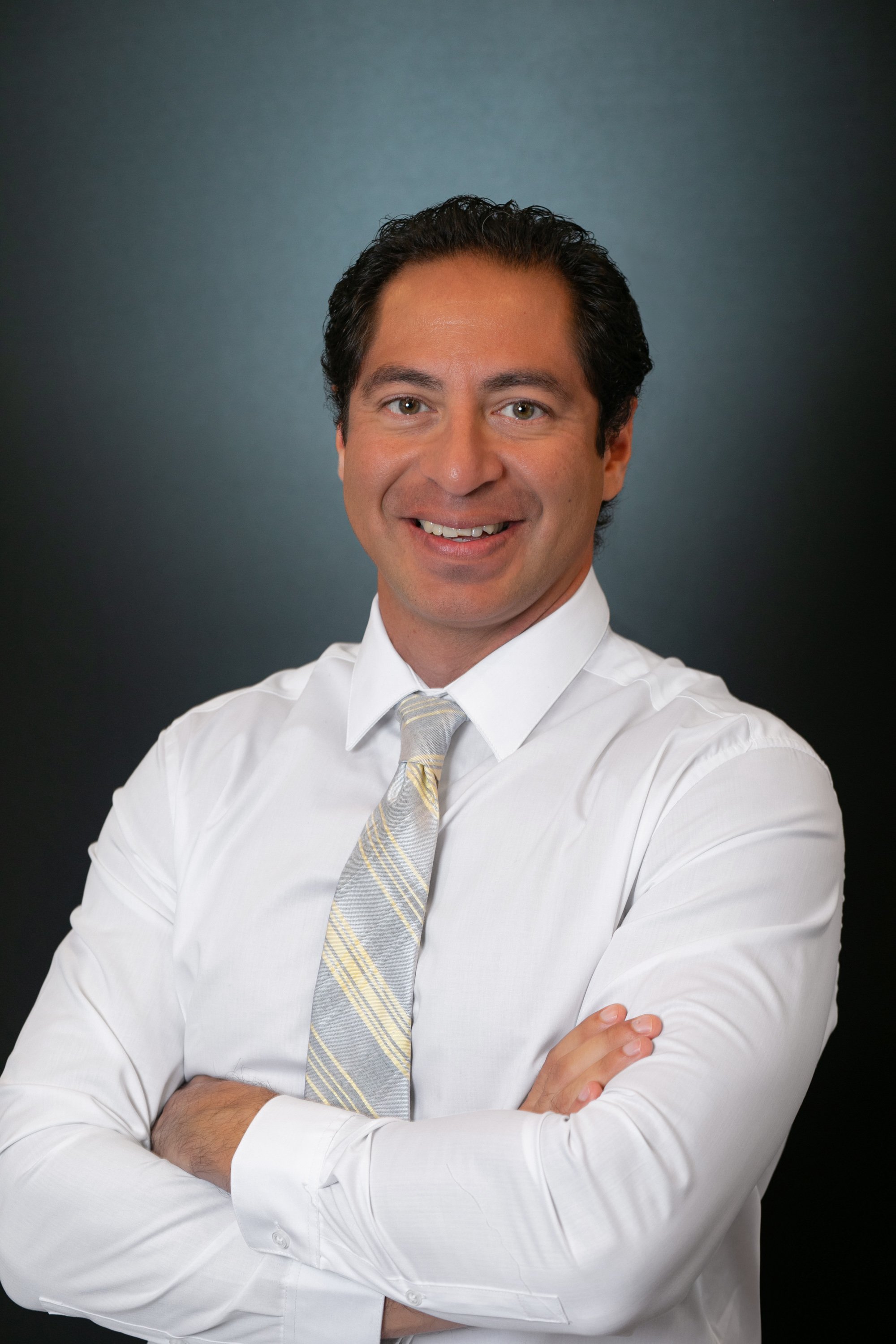 A smiling man with dark hair in a white shirt and patterned tie, standing with his arms crossed against a dark gradient background for a headshot with Cannonfire Photography in Orlando;.