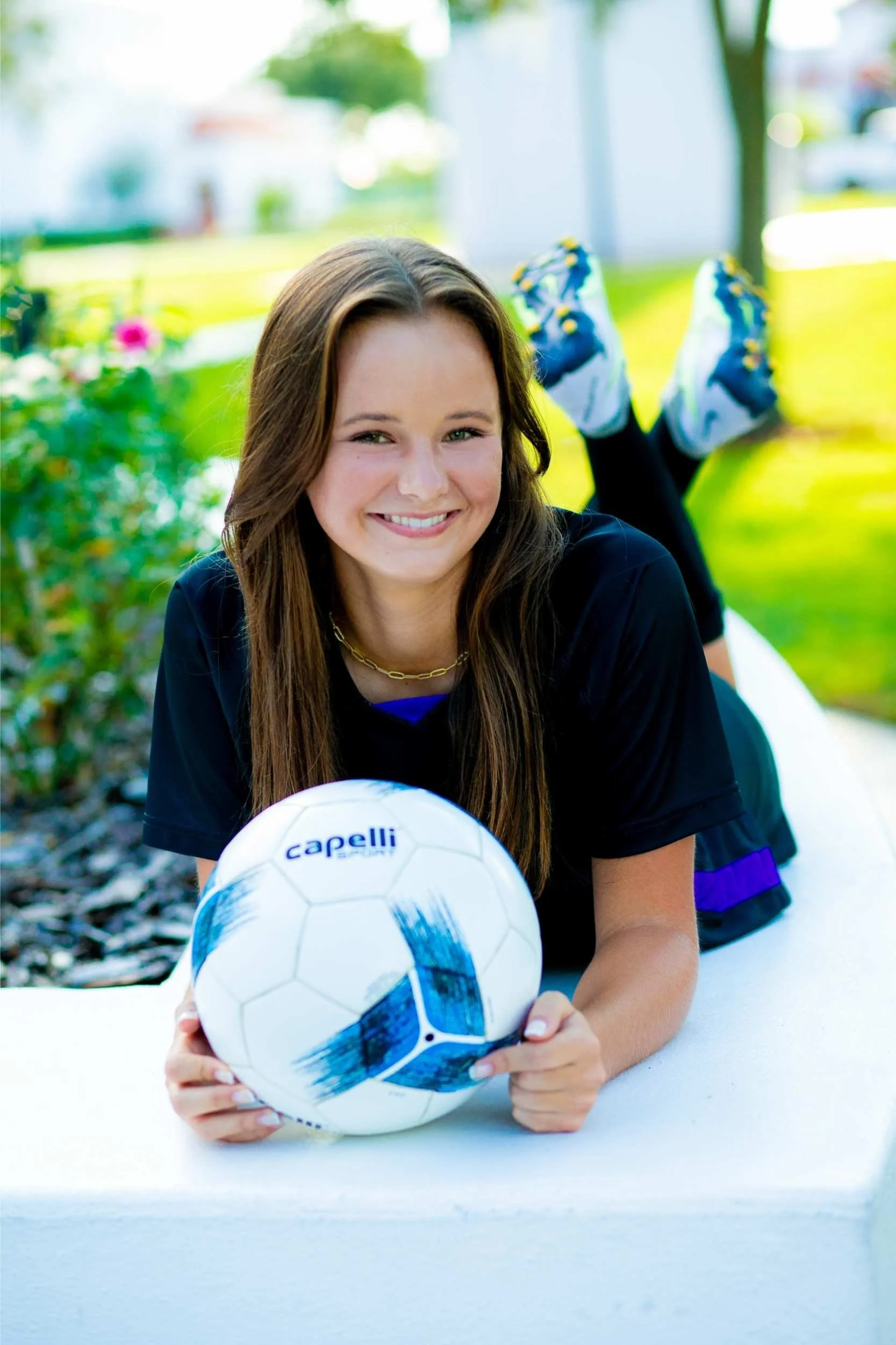 Young woman with brown hair in a black sports shirt, lying on her stomach on a white surface outdoors, holding a white soccer ball with blue accents, smiling at the camera, with blurred green grass and trees in the background.