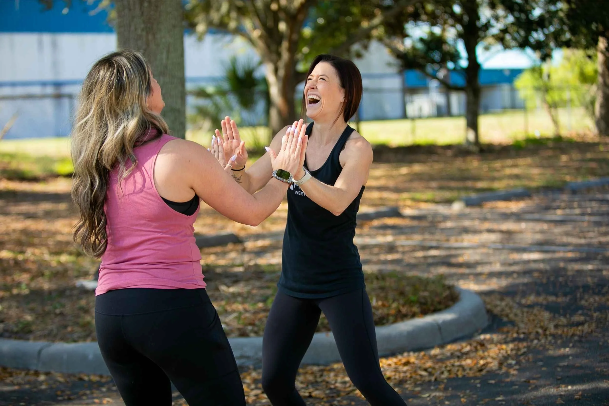 Two women are outdoors, smiling and high-fiving each other during a fitness activity in a park for an advertising photography session by Cannonfire Photography in Winter Garden.