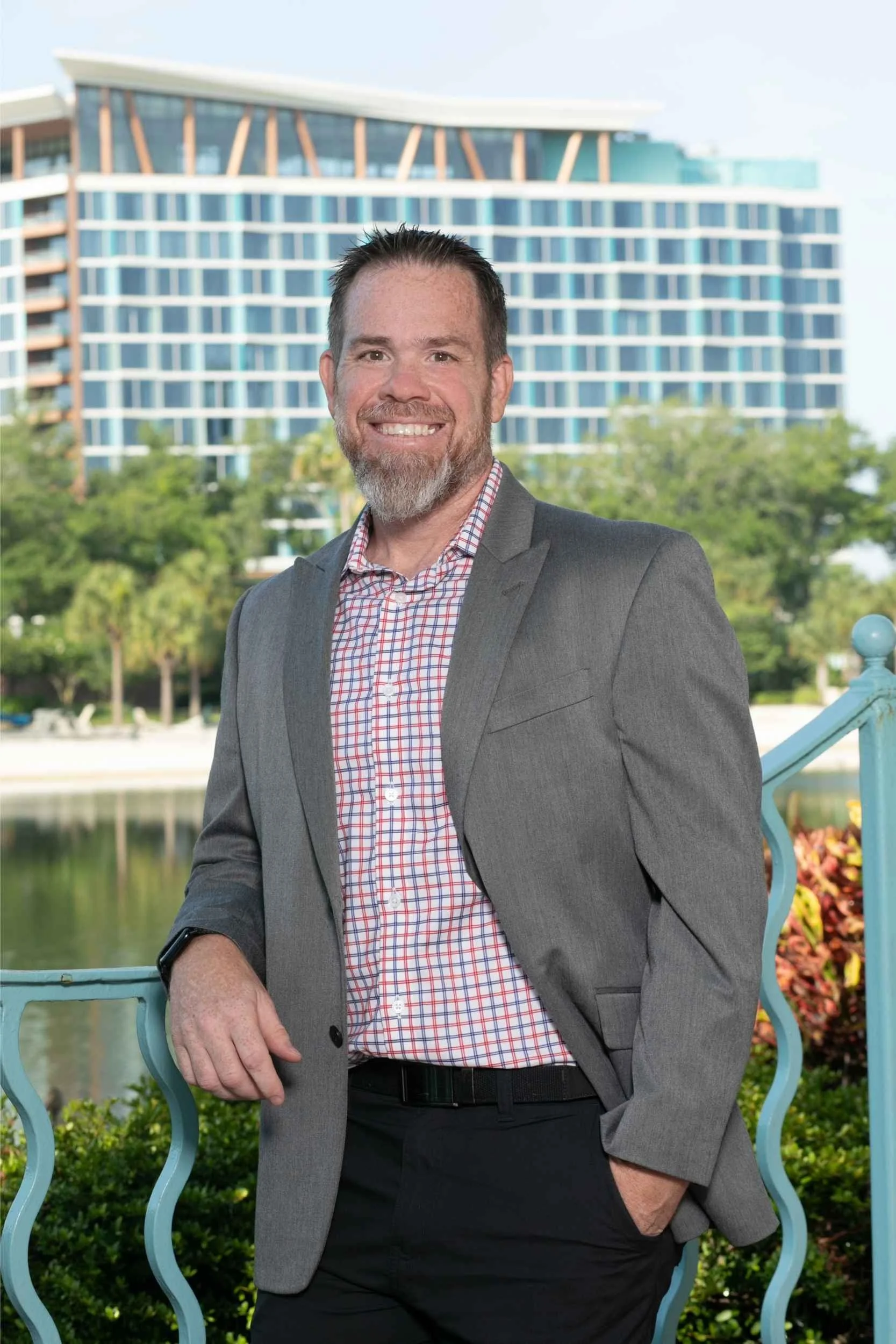 A man in a gray blazer and checkered shirt smiling outdoors with a modern building and trees in the background for a headshot with Cannonfire Photography.